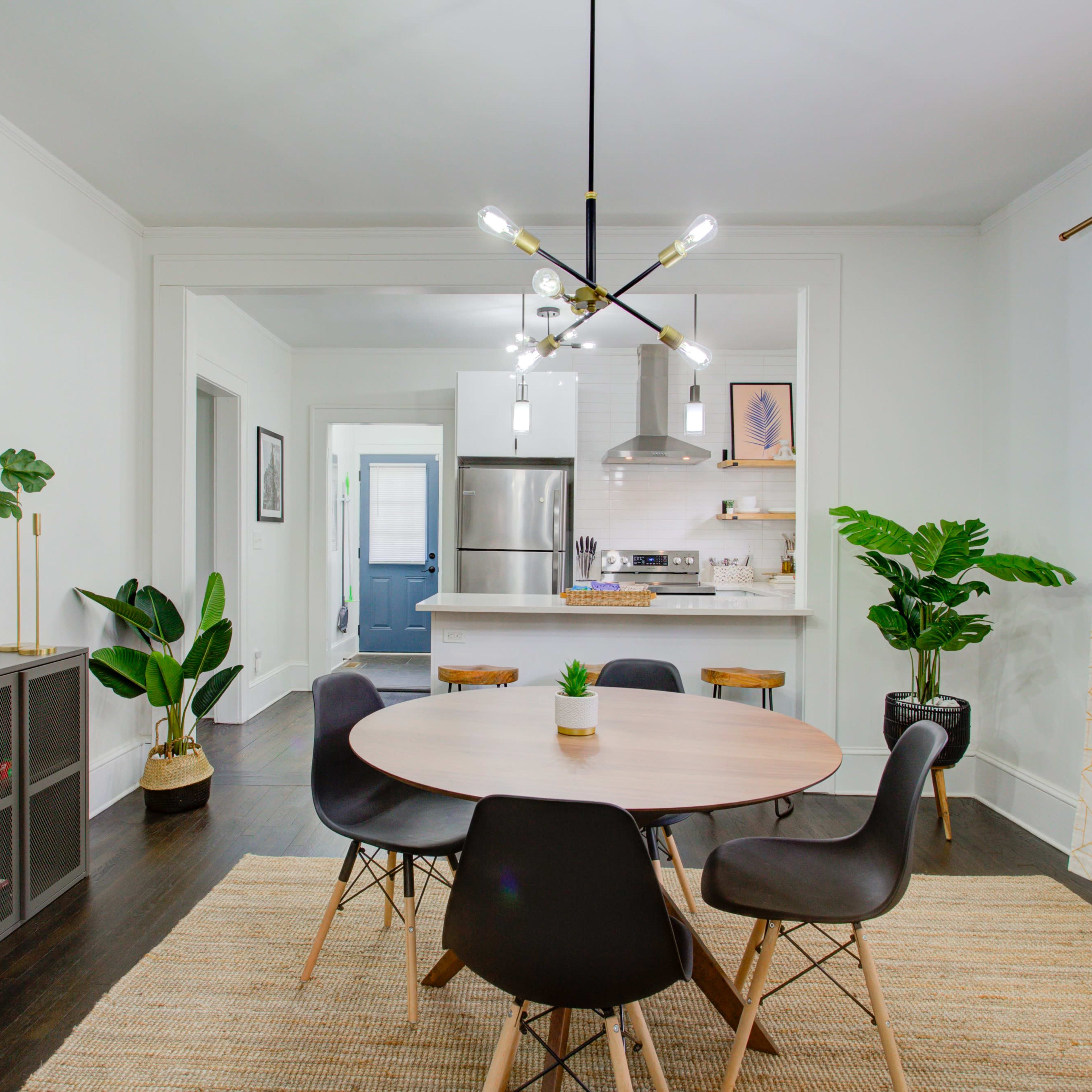 A dining area features a round table surrounded by black chairs, complemented by a wall of plants and a modern light fixture, with an open view into a kitchen.