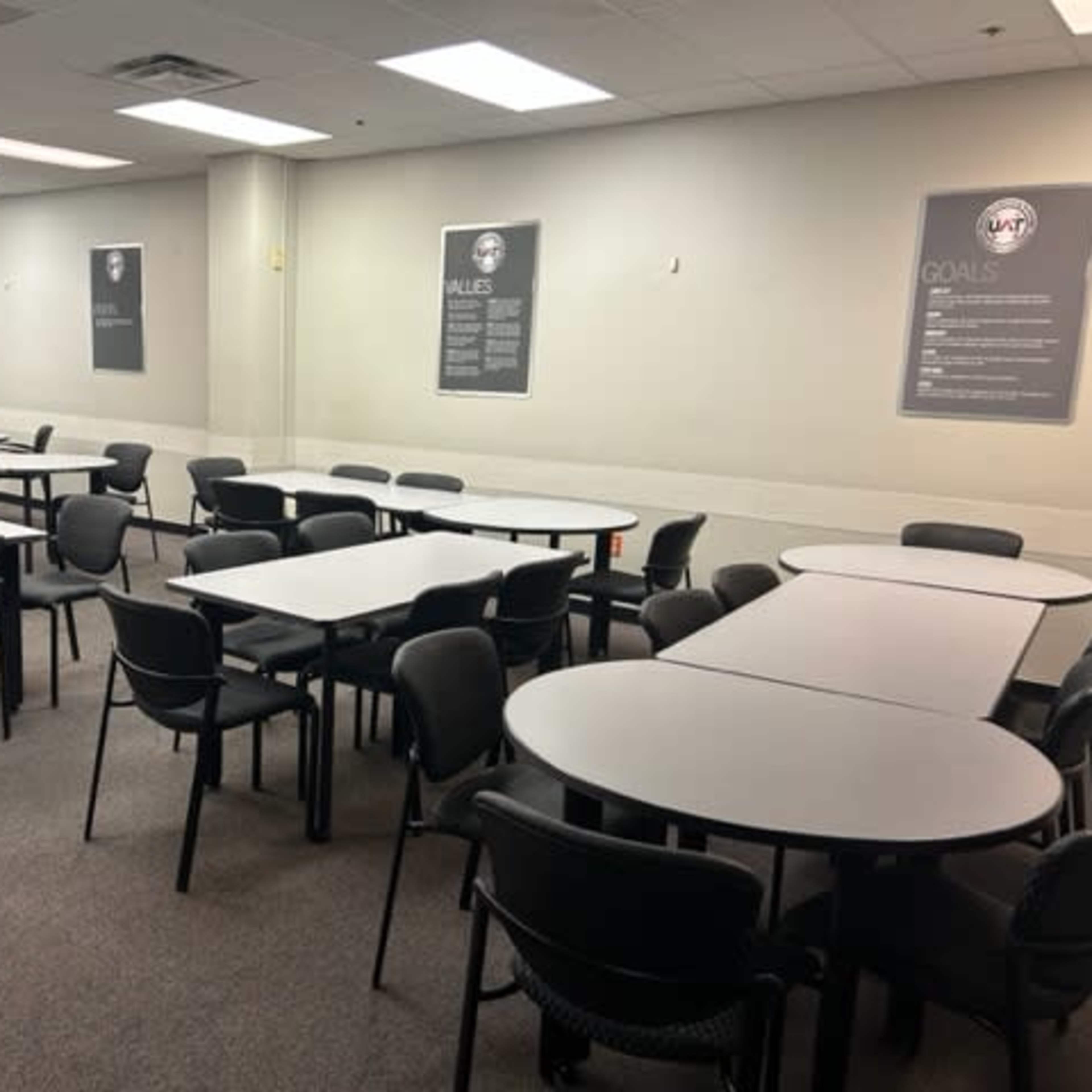 A conference room with several tables and chairs arranged throughout, and posters displayed on the beige walls.