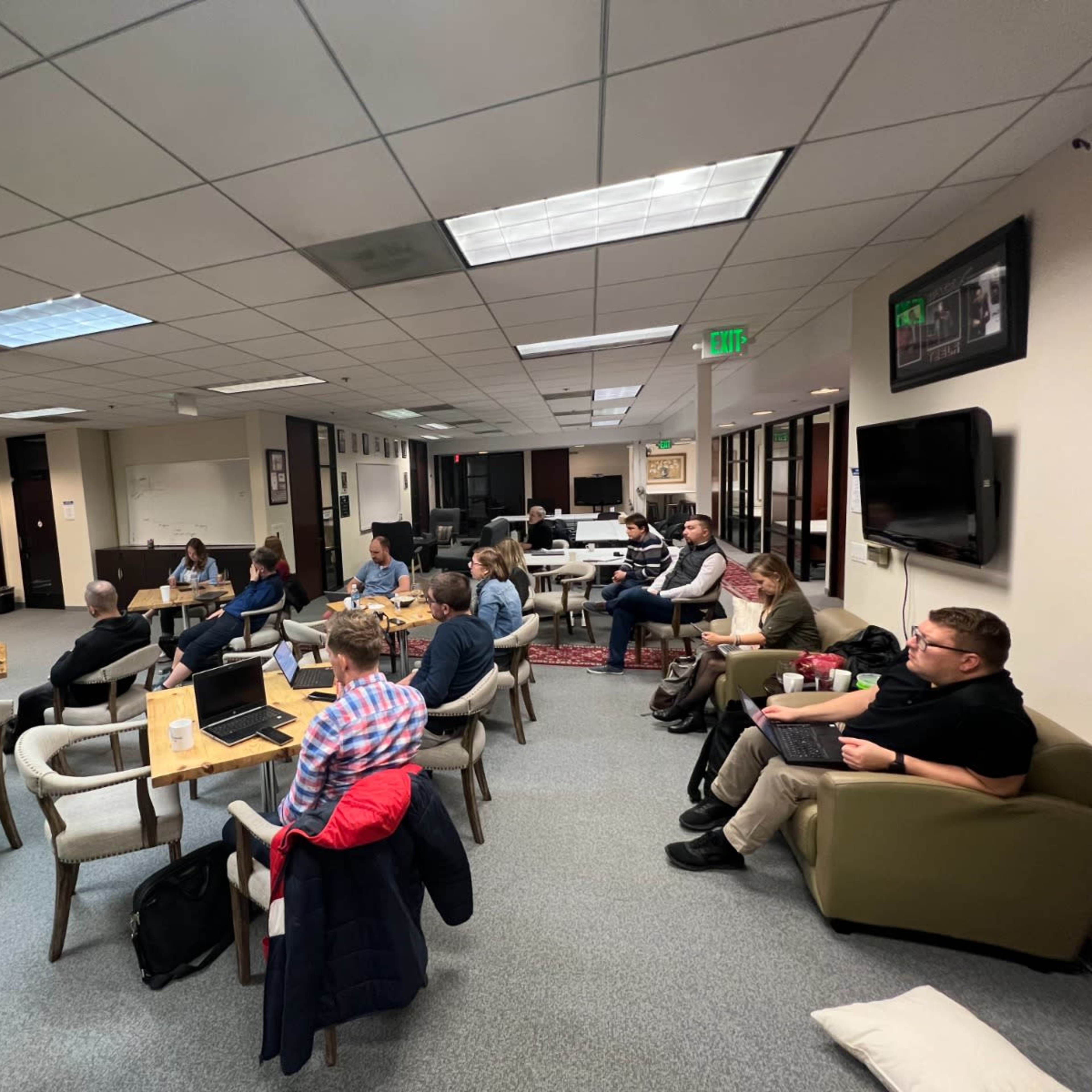 A group of people sits at tables and on chairs in a well-lit room, actively working on laptops and engaging in discussion.
