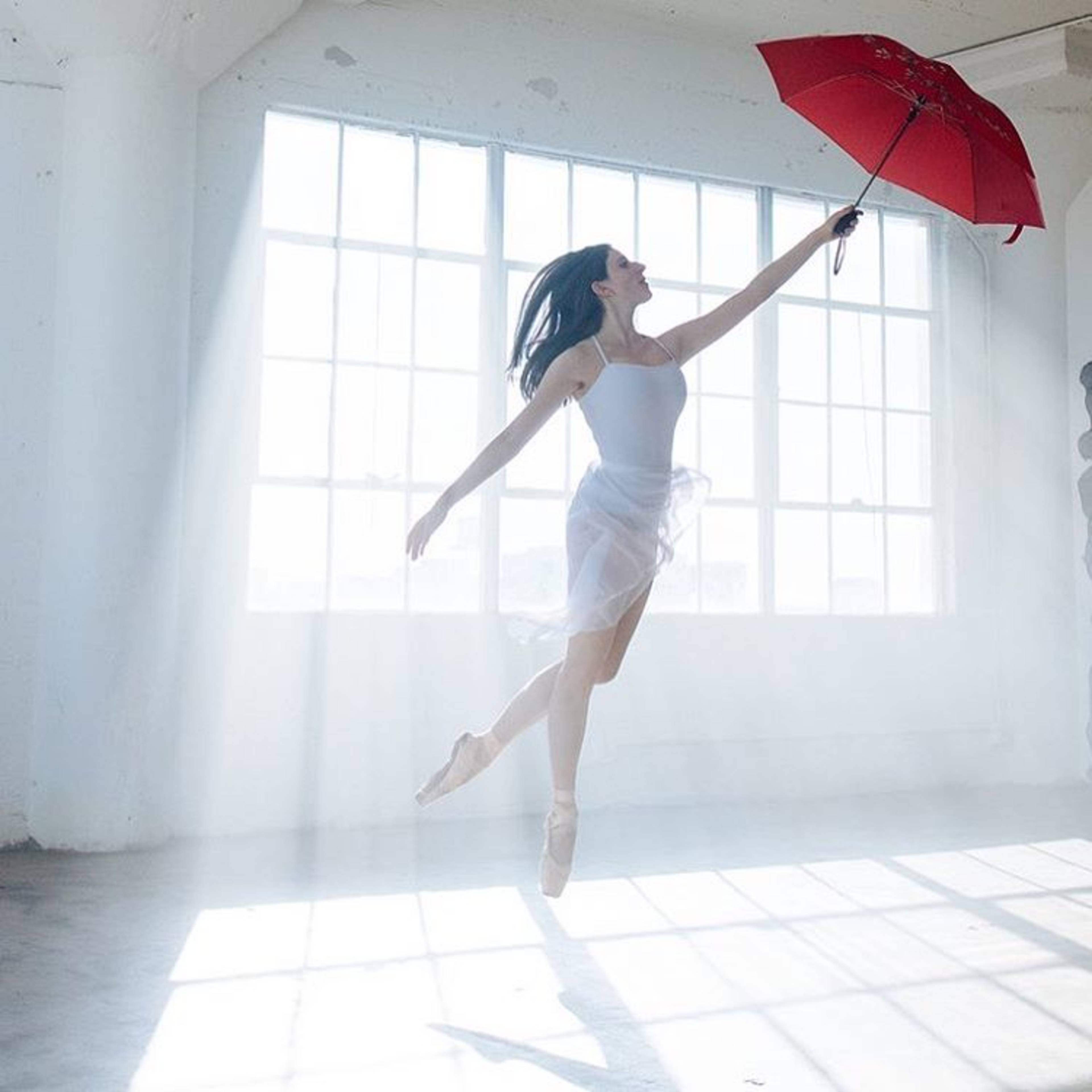 A dancer jumps while holding a red umbrella in a sunlit room with large windows.
