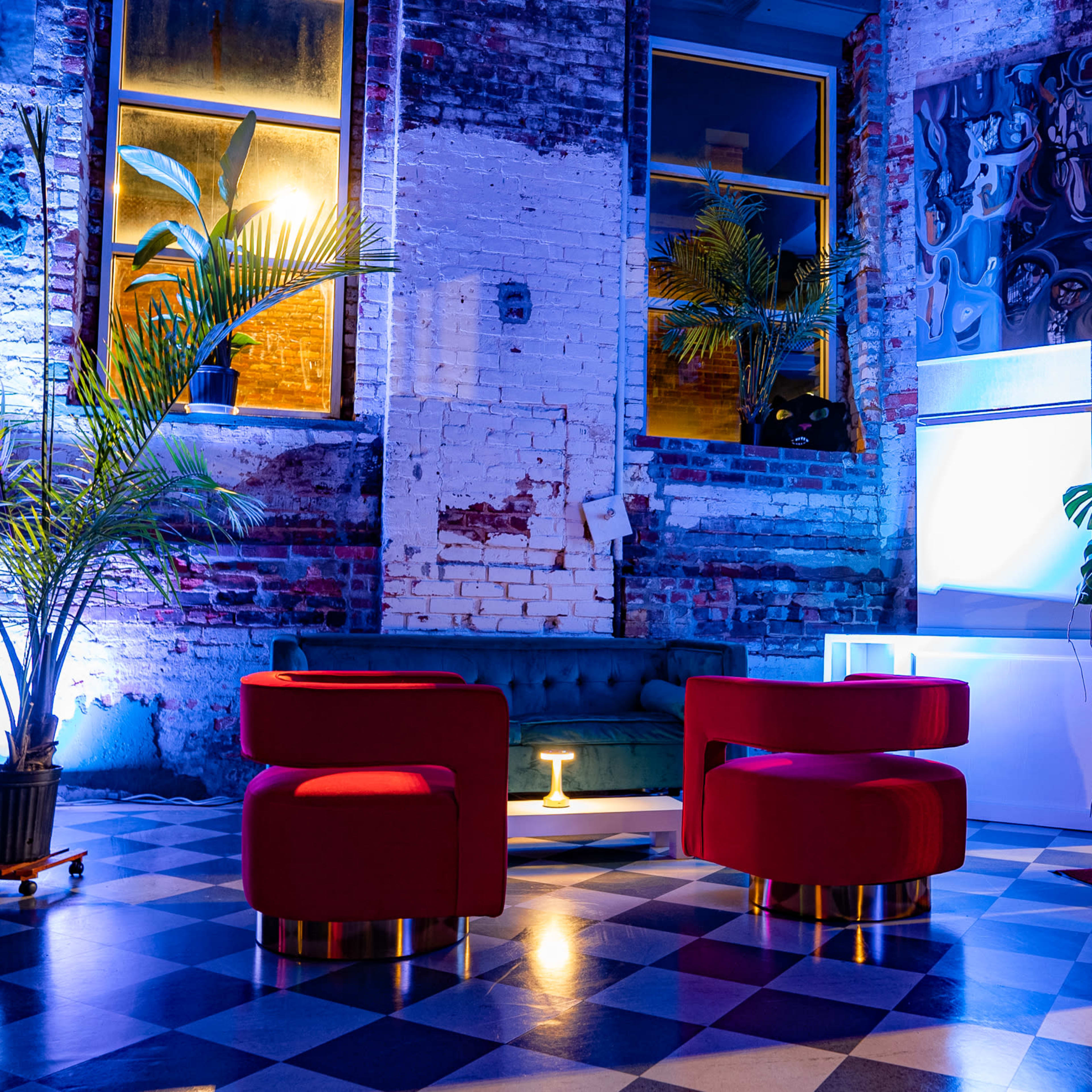 A modern lounge area with a green couch and two red, circular chairs on a checkered floor, surrounded by potted plants and illuminated by colorful lighting in a rustic brick setting.