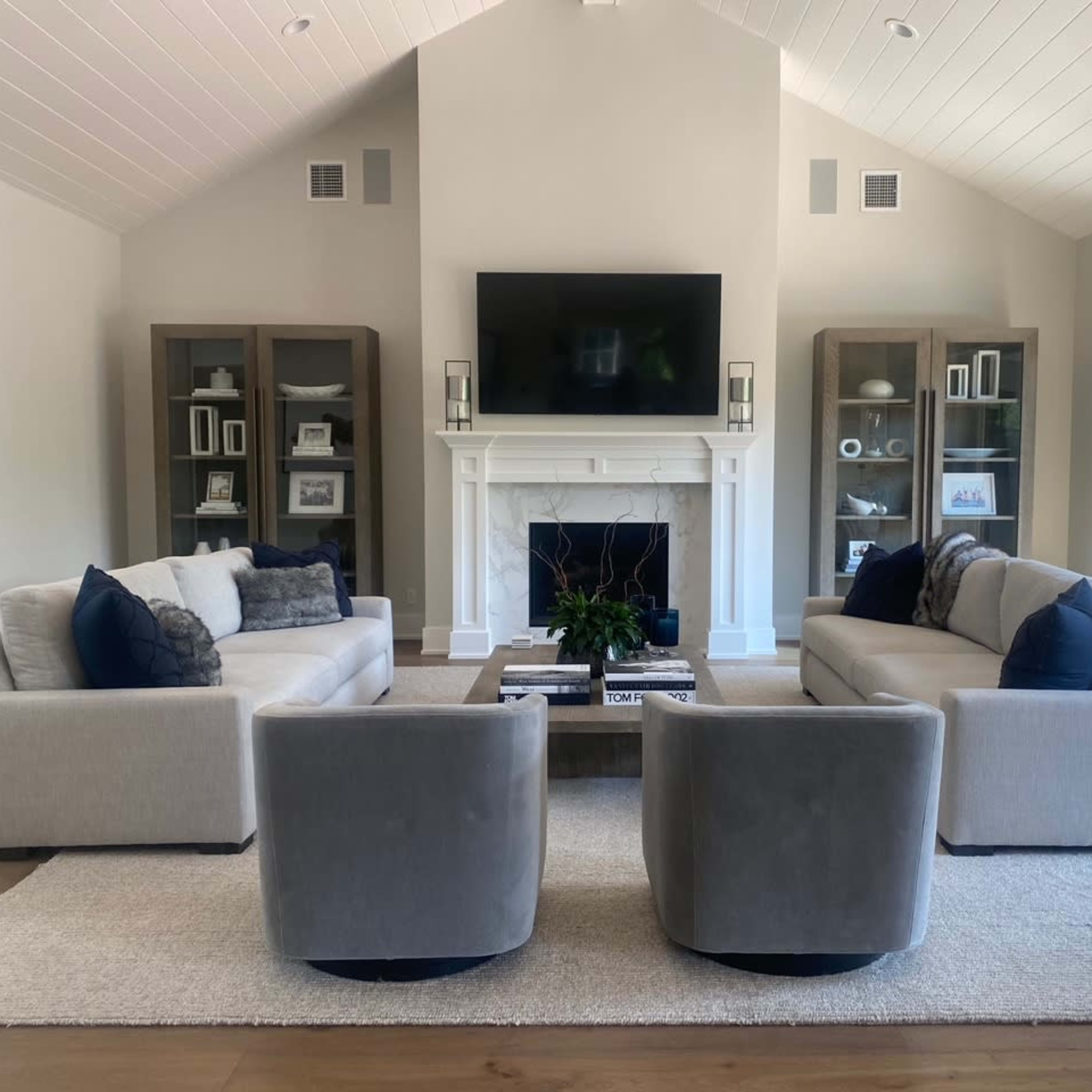 A modern living room featuring two gray sofas and two matching armchairs arranged around a central coffee table, with a television mounted above a white fireplace.