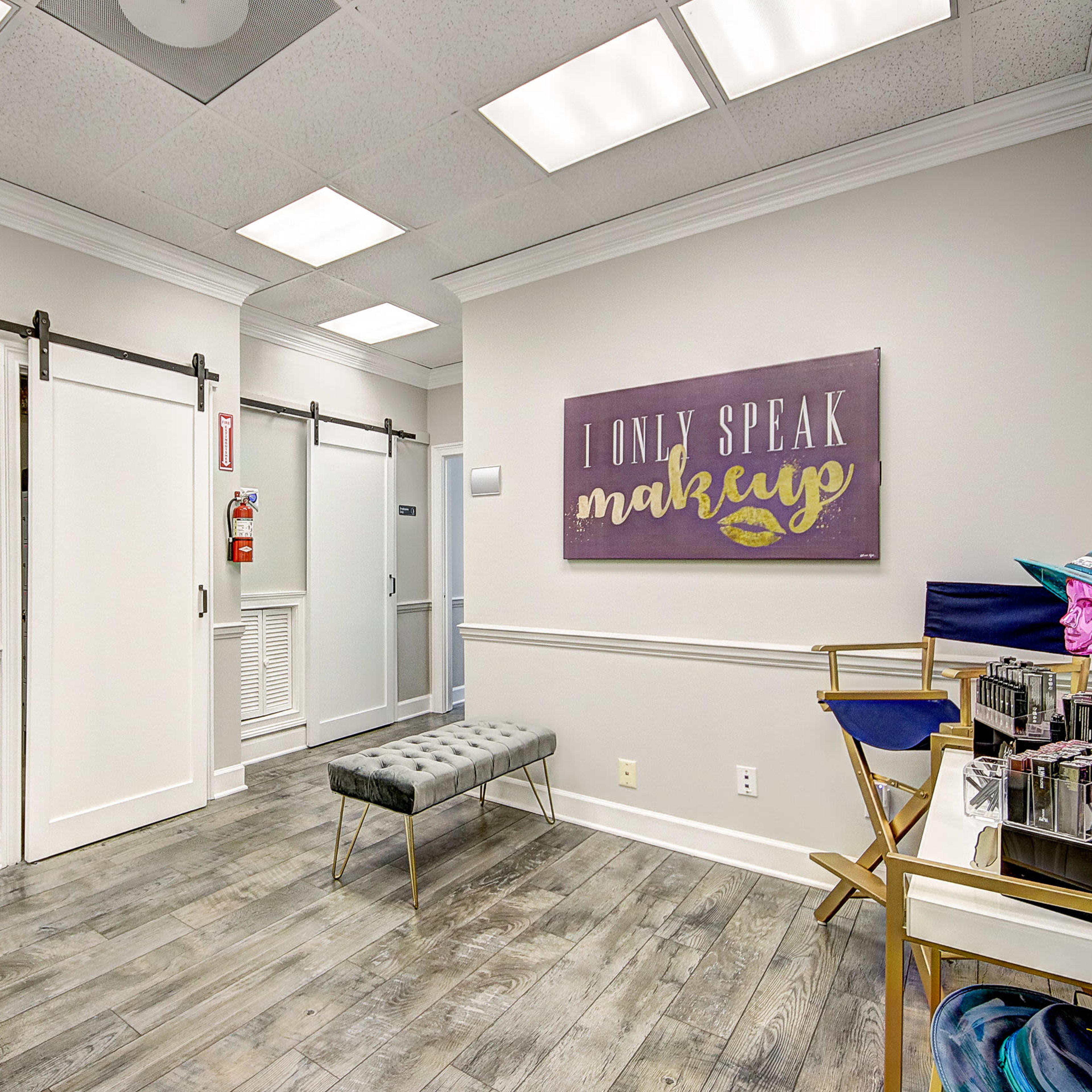 A well-lit interior space with a wooden floor, featuring a bench, a large mirror, and a sign that reads "I ONLY SPEAK MAKEUP" on the wall.