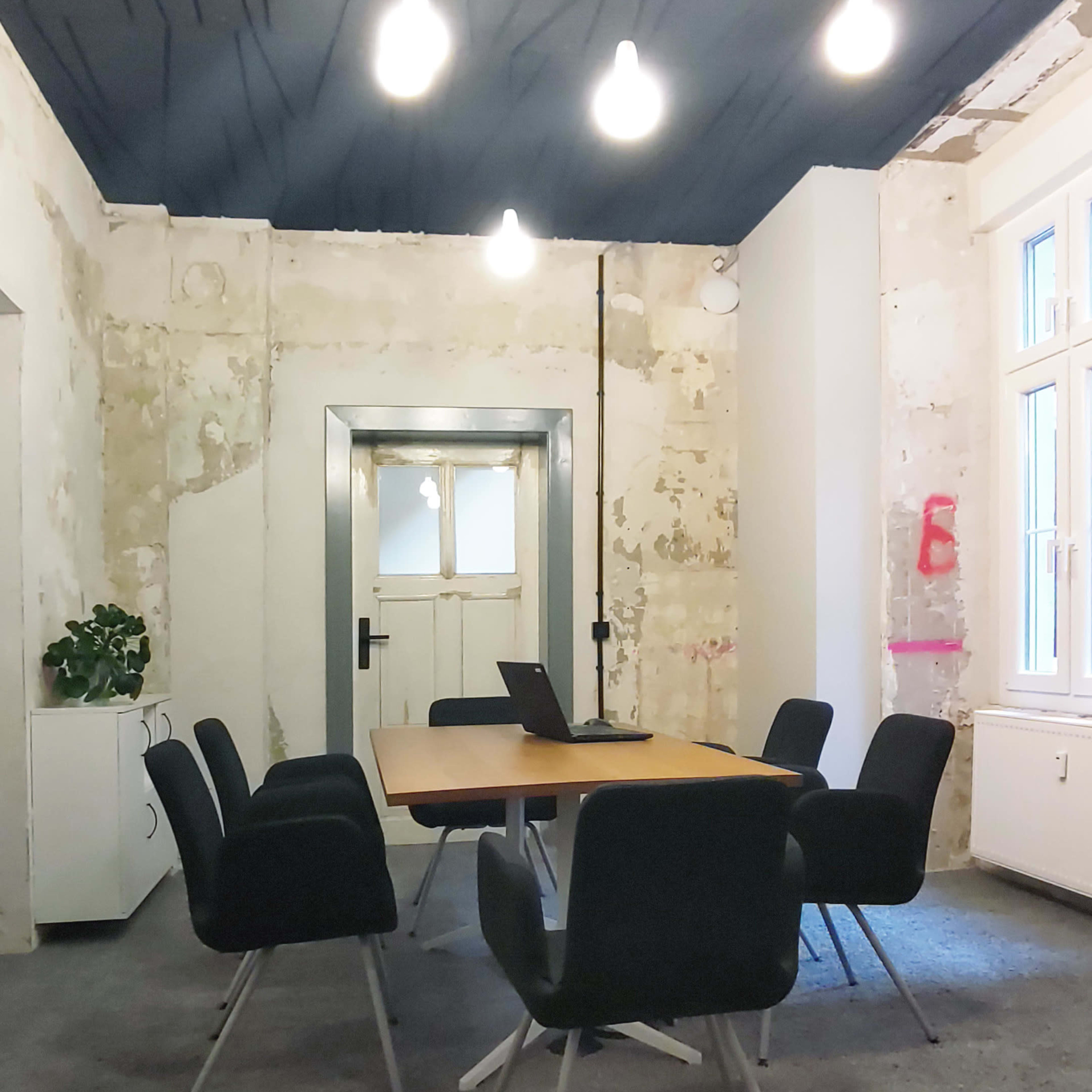 A conference room features a central wooden table surrounded by six chairs, with a laptop on the table and exposed light bulbs hanging from the ceiling.
