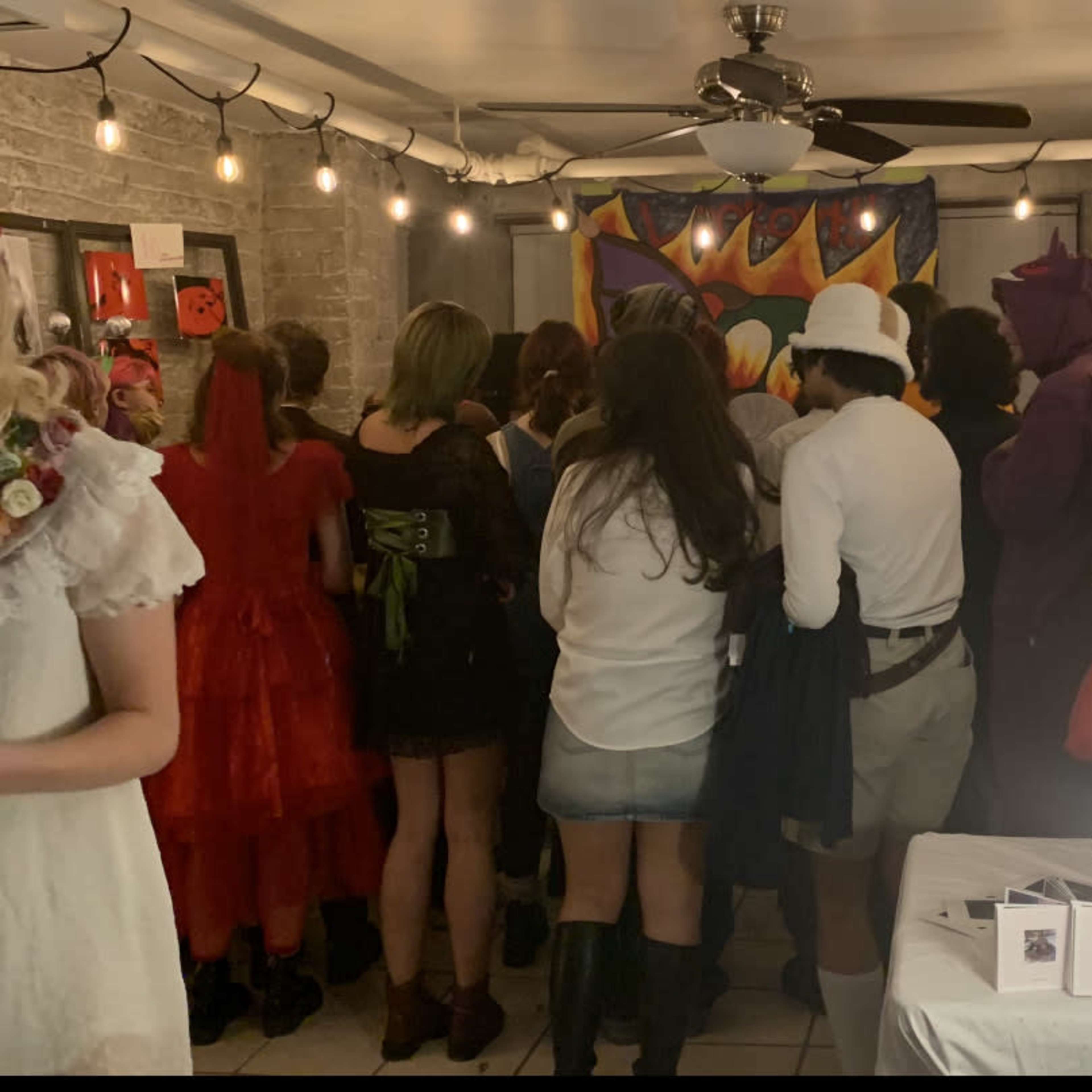 A group of people in diverse costumes gathers around a colorful mural in a dimly lit room.
