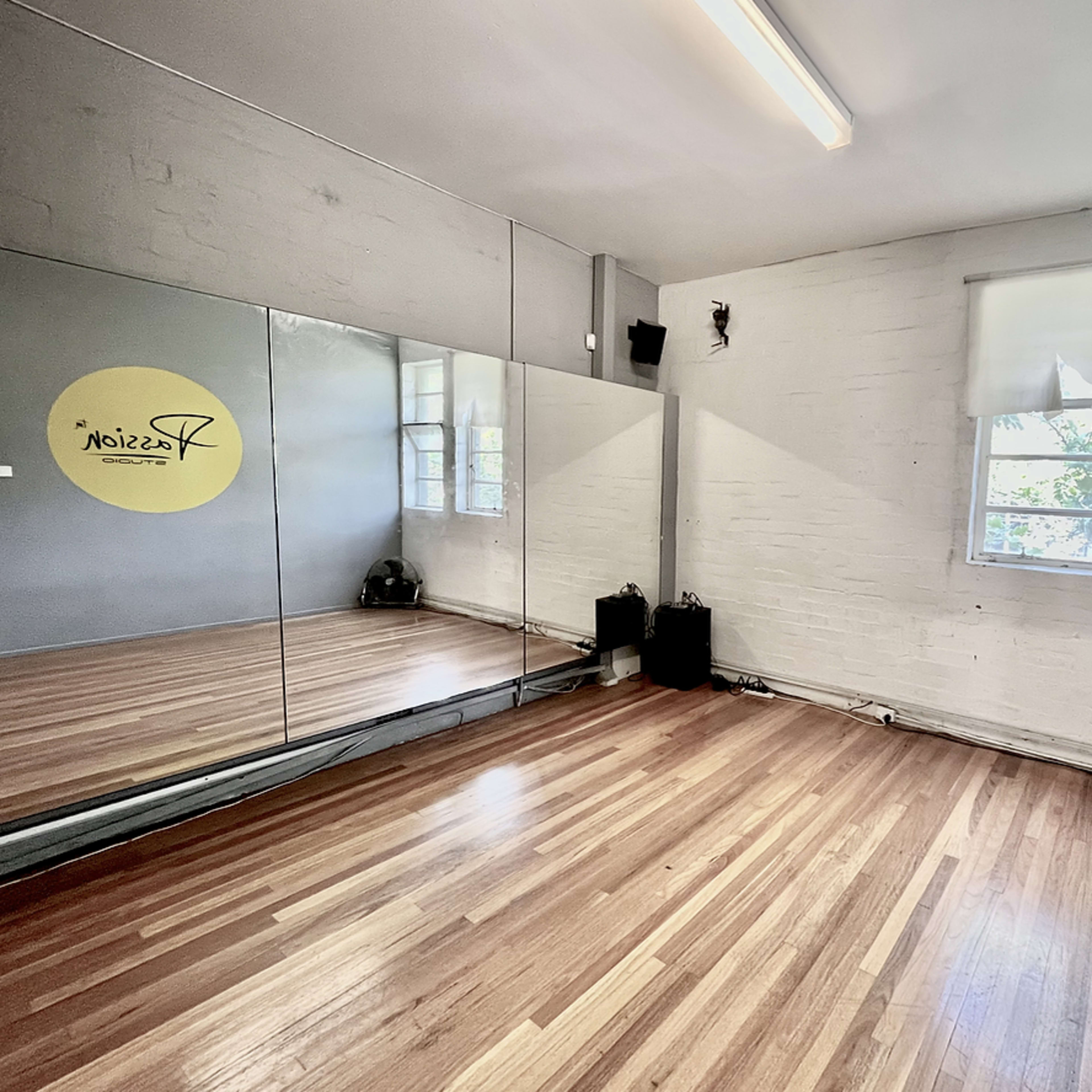 A mirrored wall reflects a bright dance studio with wooden flooring and large windows.