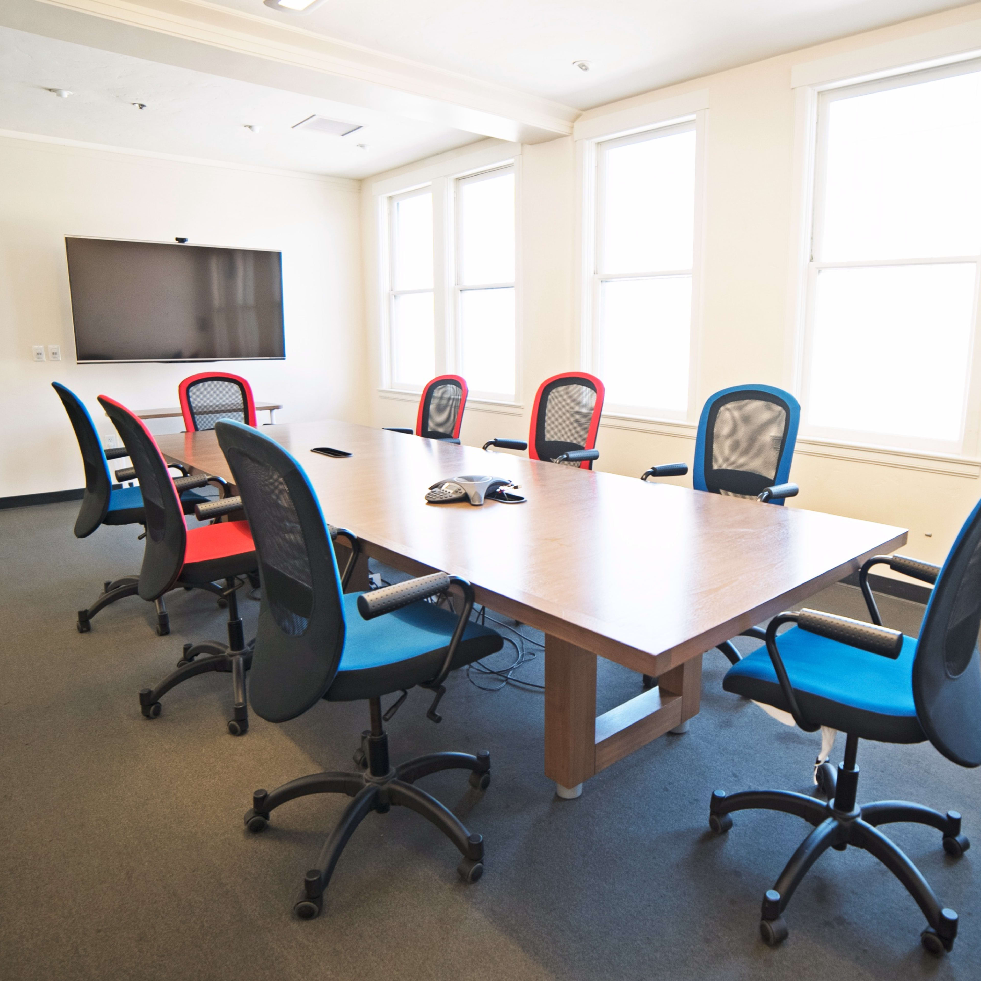 A conference room features a large wooden table surrounded by six office chairs in red and blue, with a television mounted on the wall and windows allowing natural light.