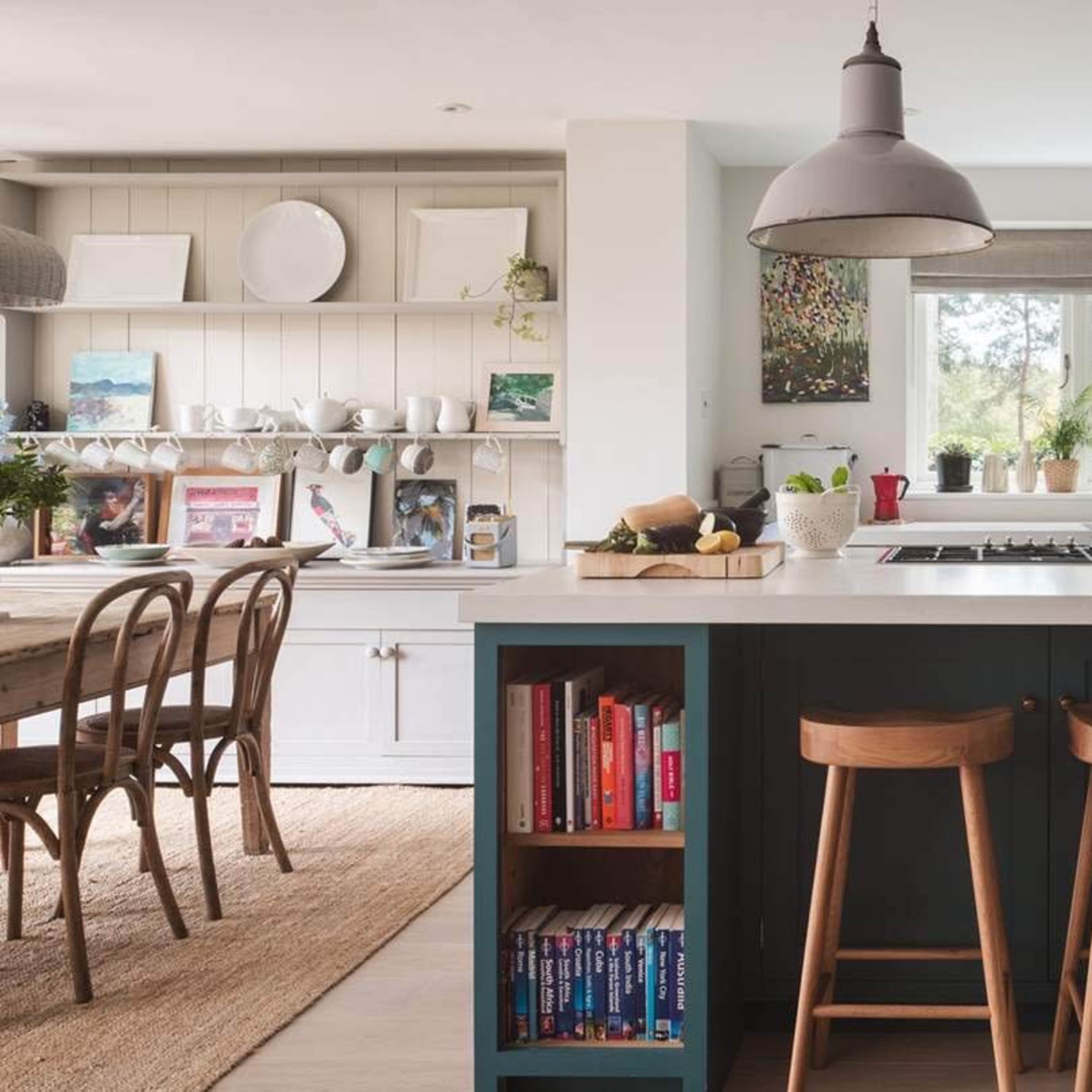 A modern kitchen features a central island with a stovetop, two wooden stools, and bookshelves underneath, while a dining area is visible in the background.