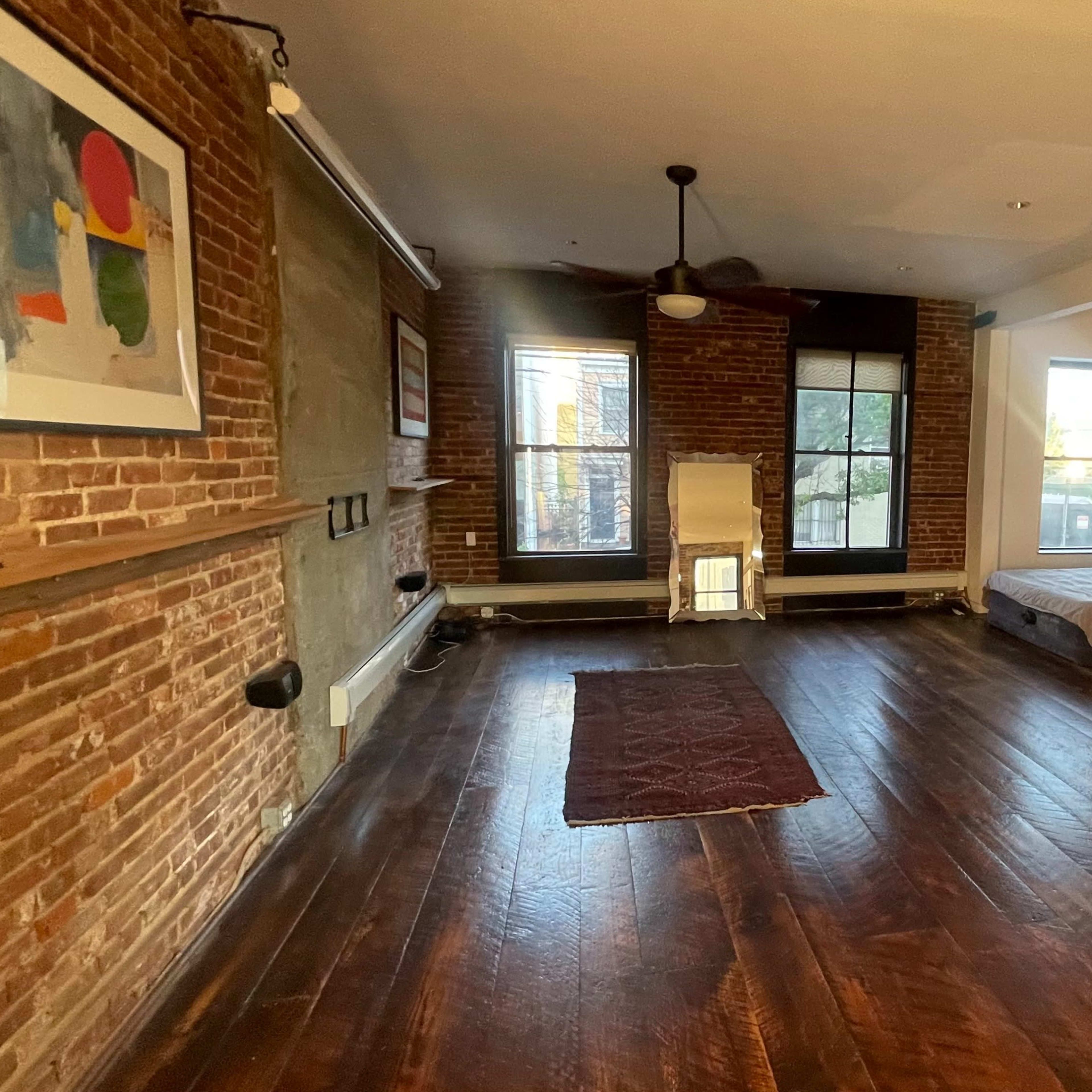 A spacious room with exposed brick walls, hardwood flooring, a mirror, a bed in the corner, and large windows allowing light to enter.