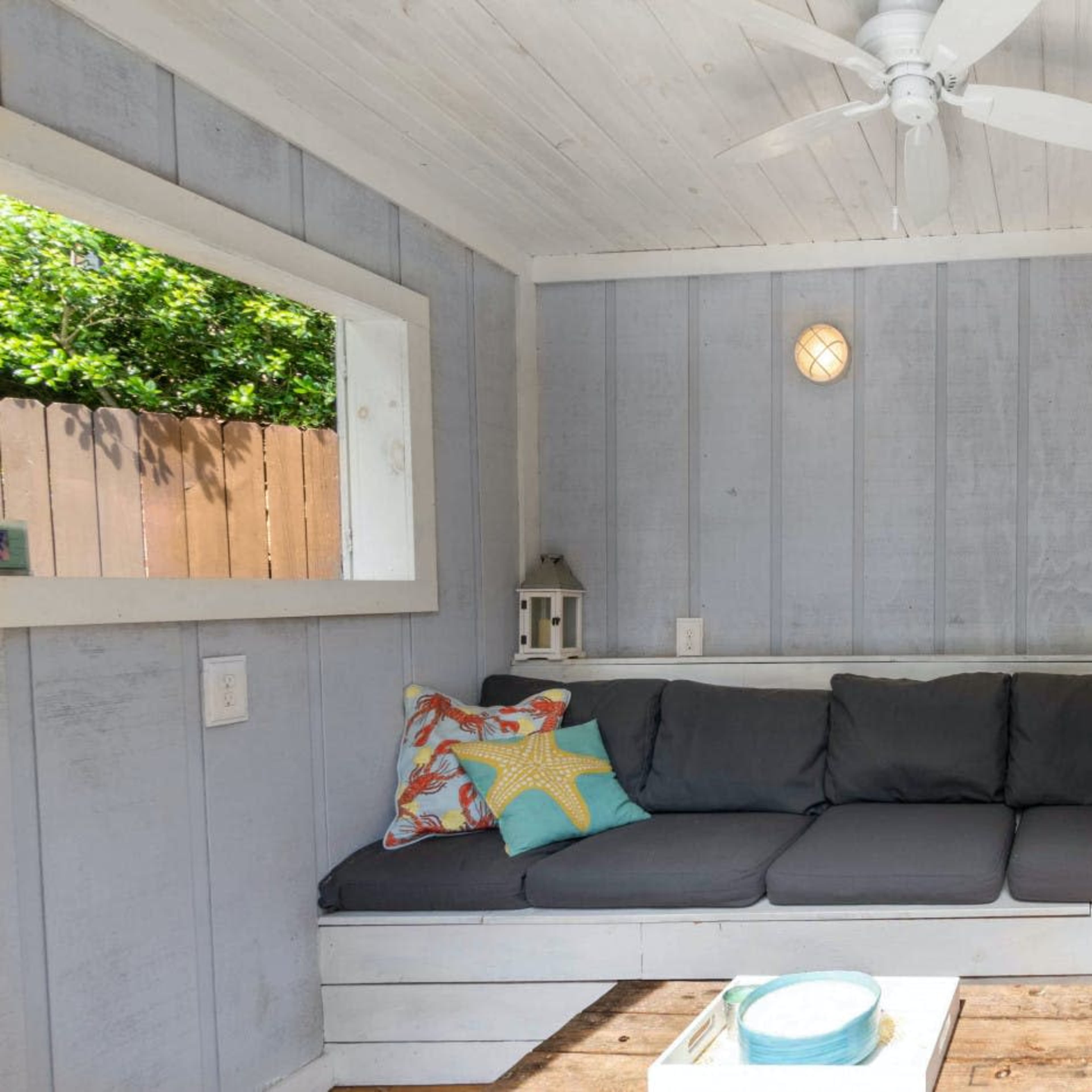 A cozy seating area with a gray sectional couch, colorful pillows, a central table, and a window allowing natural light.