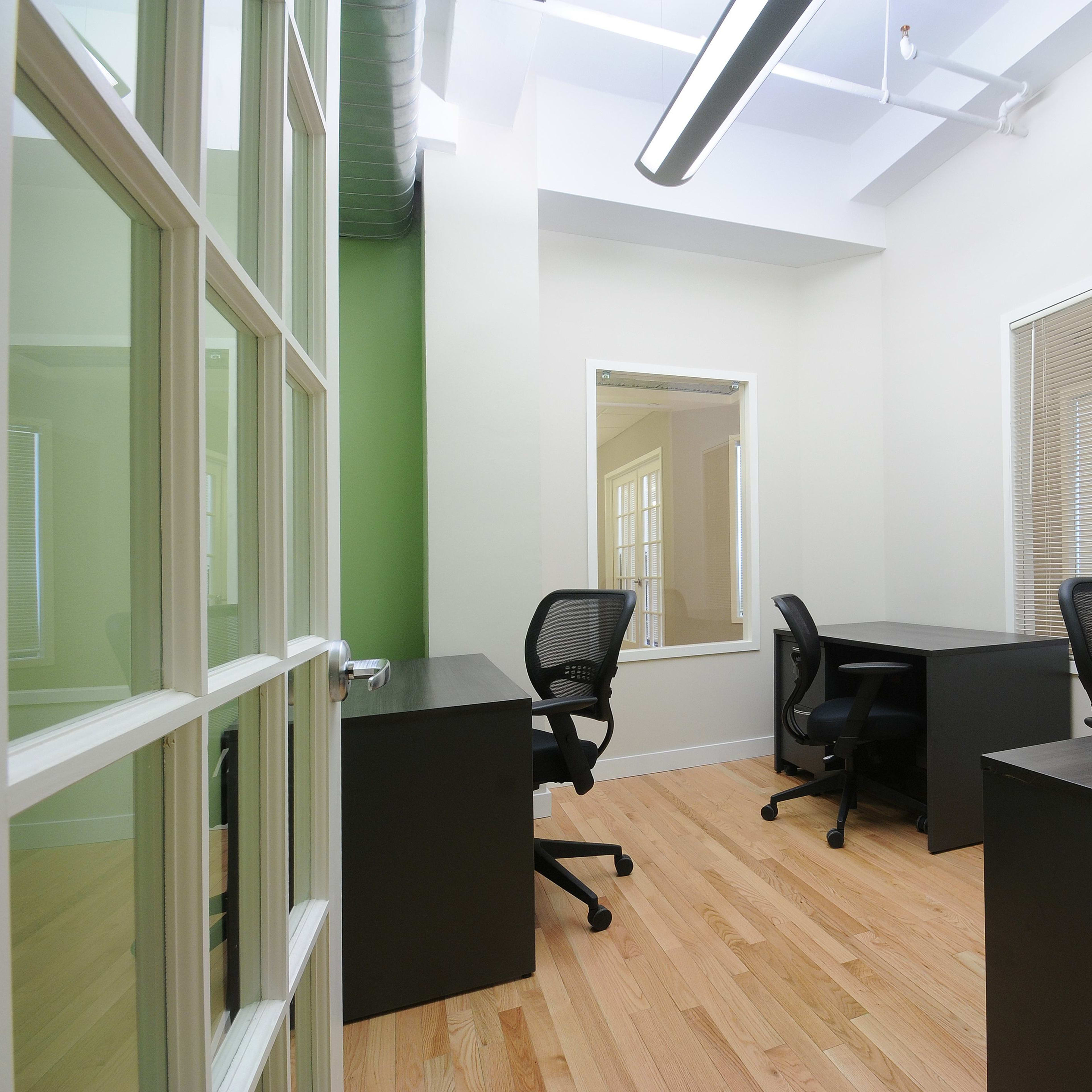 A modern office space with two black desks and rolling chairs, featuring light hardwood floors and green accent walls.