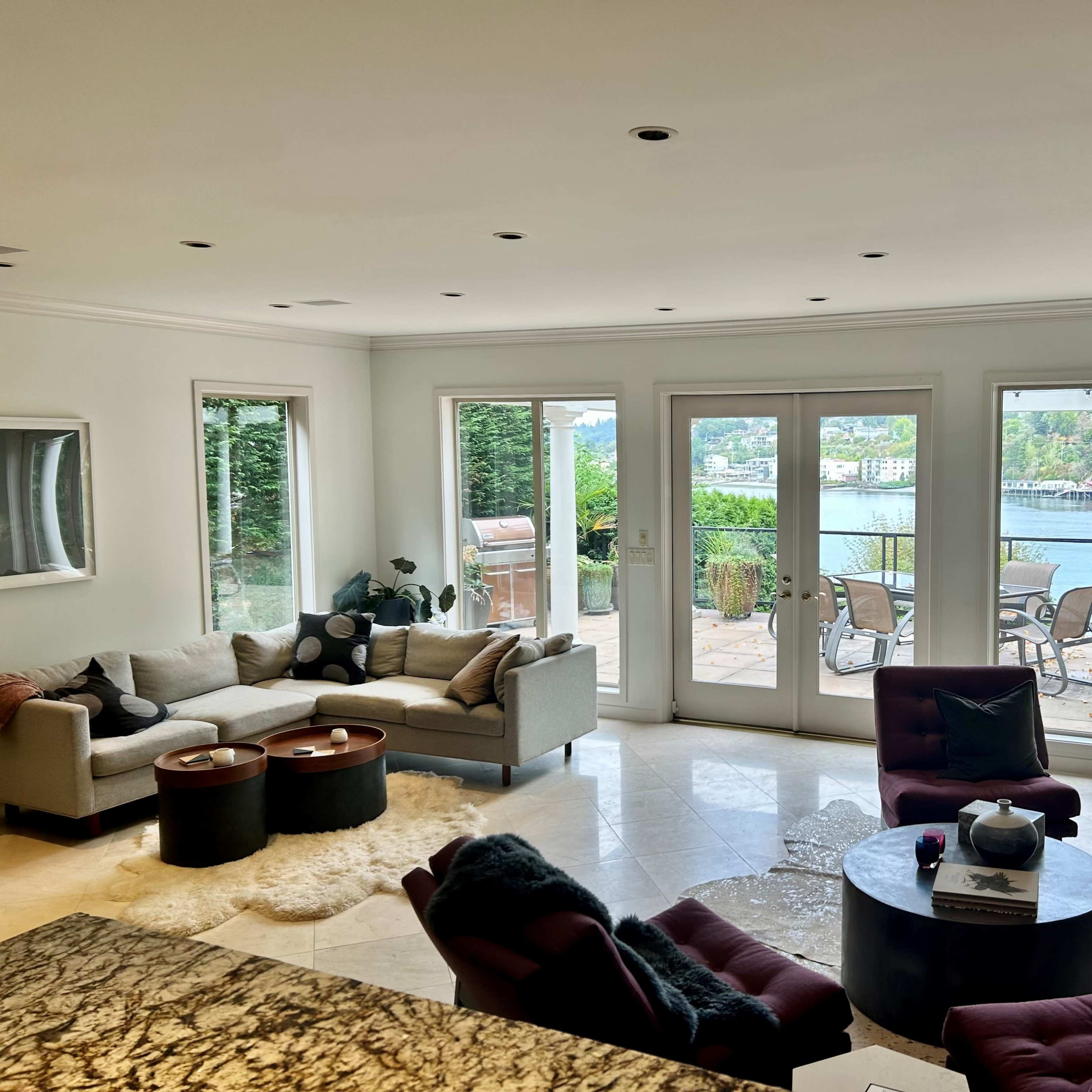 A well-lit living room features a beige sectional sofa, two maroon chairs, and large glass doors showcasing a view of the water and patio area.