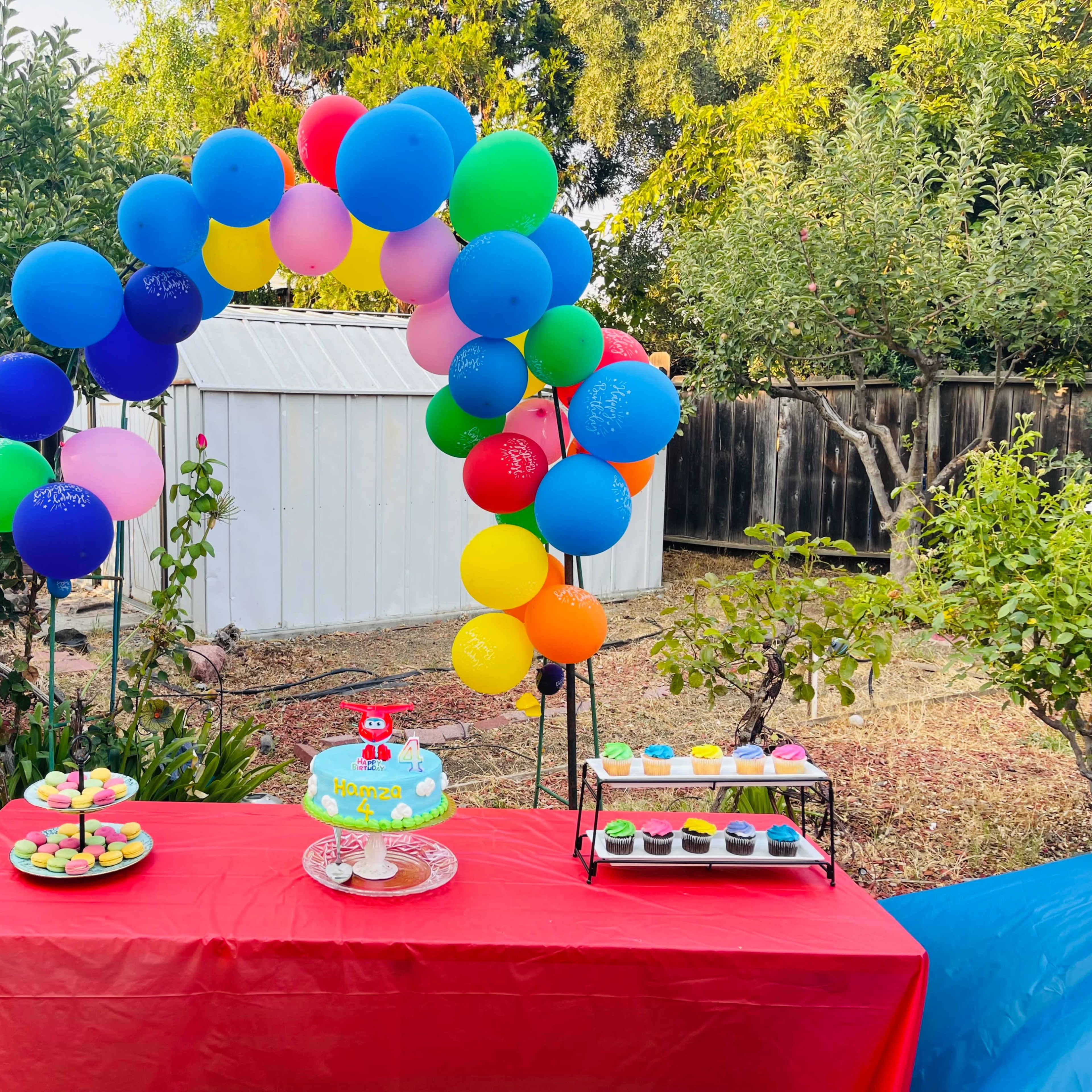 A festive outdoor table is decorated with a colorful balloon arch, featuring a cake and assorted cupcakes alongside a platter of cookies.