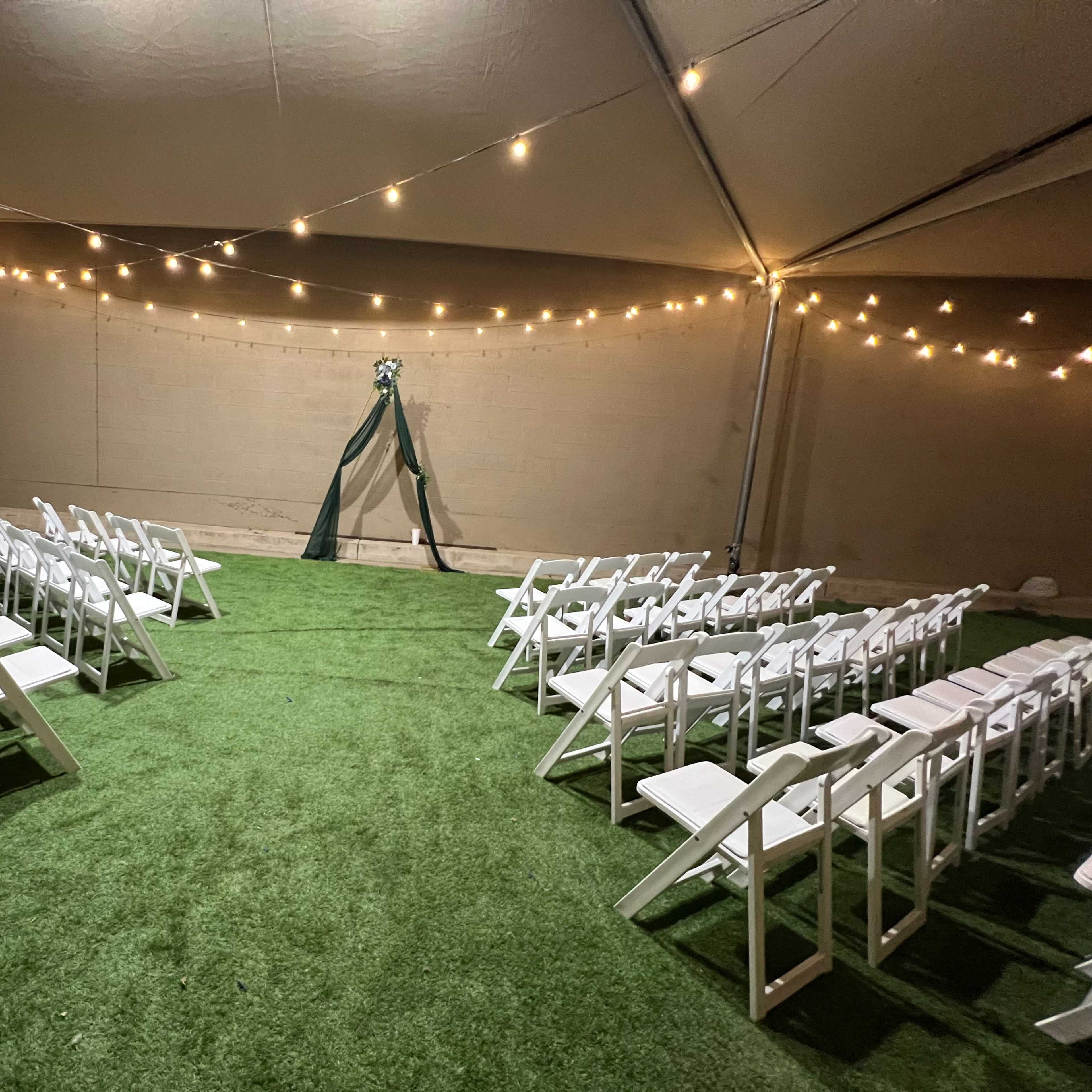 A tented wedding setup features neatly arranged white chairs on artificial grass facing a decorated arch under string lights.