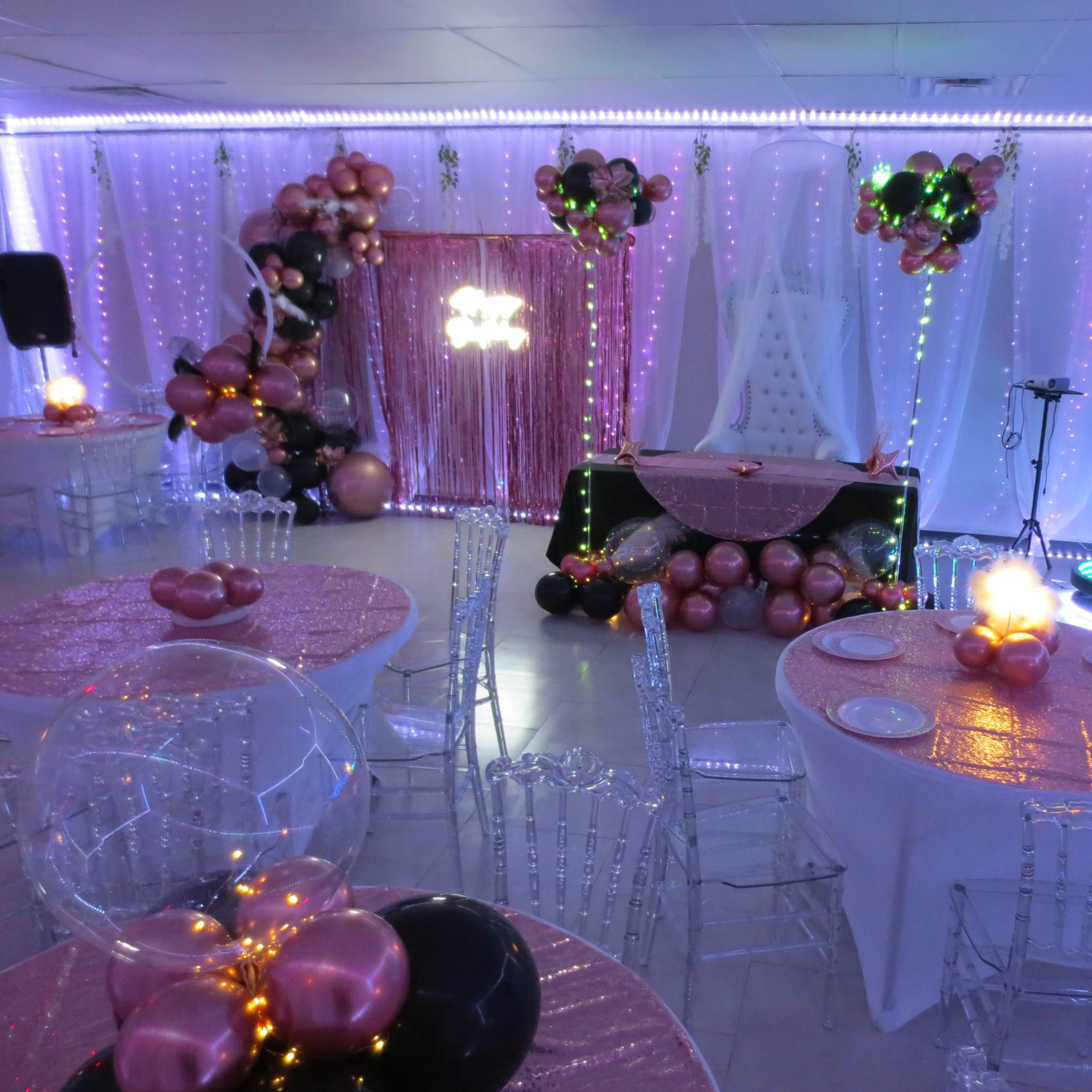 A decorated event space featuring round tables with crystal chairs, adorned with pink and black balloons, and a backdrop of lights and decorations at the front.