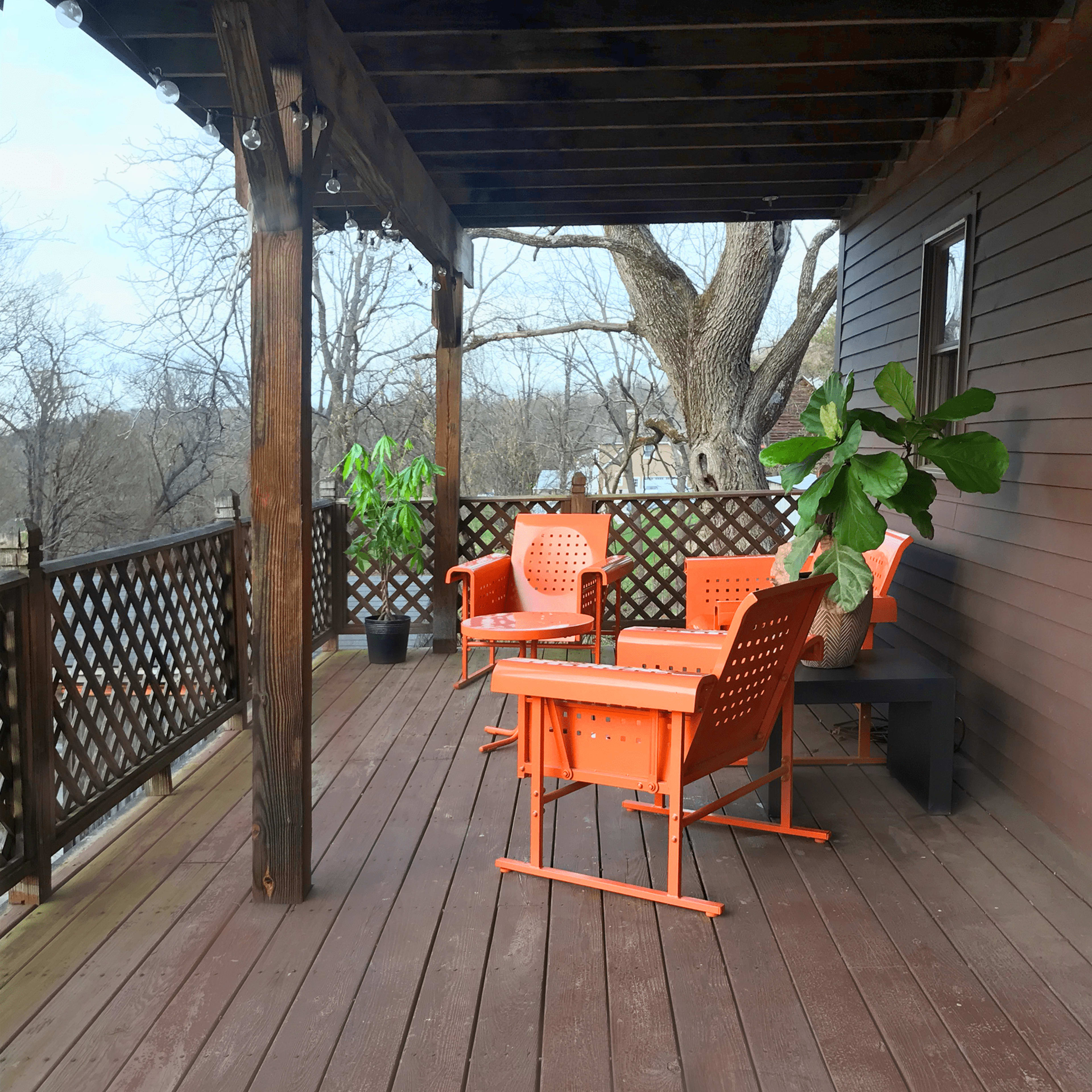 A wooden deck with orange patio chairs and a small table, surrounded by a railing and trees in the background.