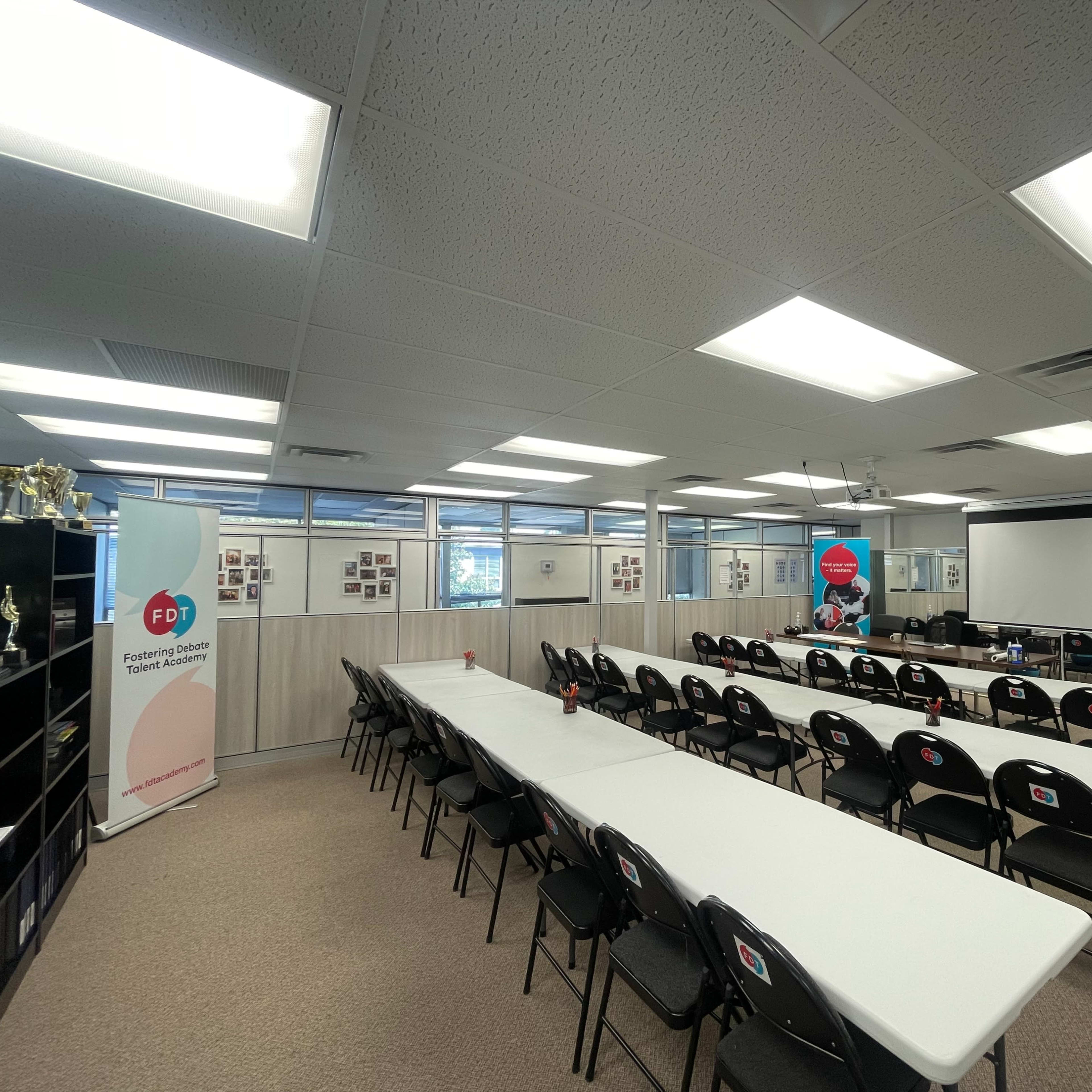 A clean conference room arranged with rows of white tables and black chairs, equipped with a projector screen and presentation tools.