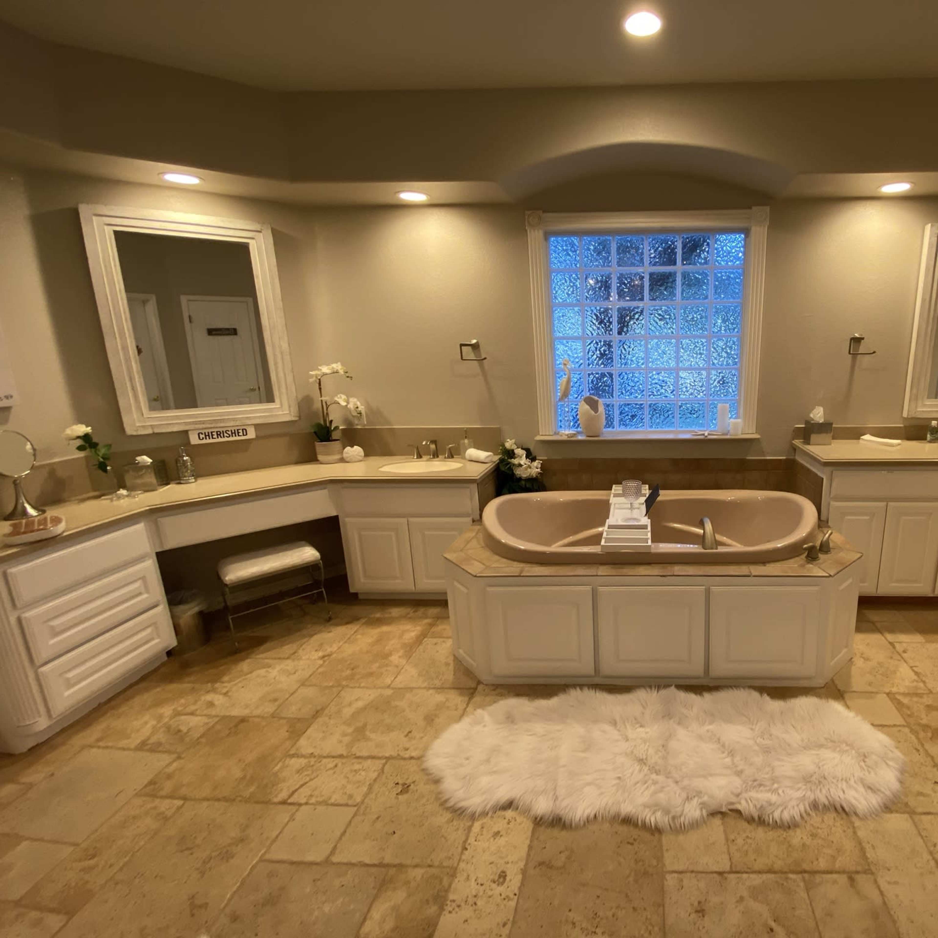 A spacious bathroom features a central bathtub with a tiled surround, dual sinks, and large mirrors illuminated by soft lighting.
