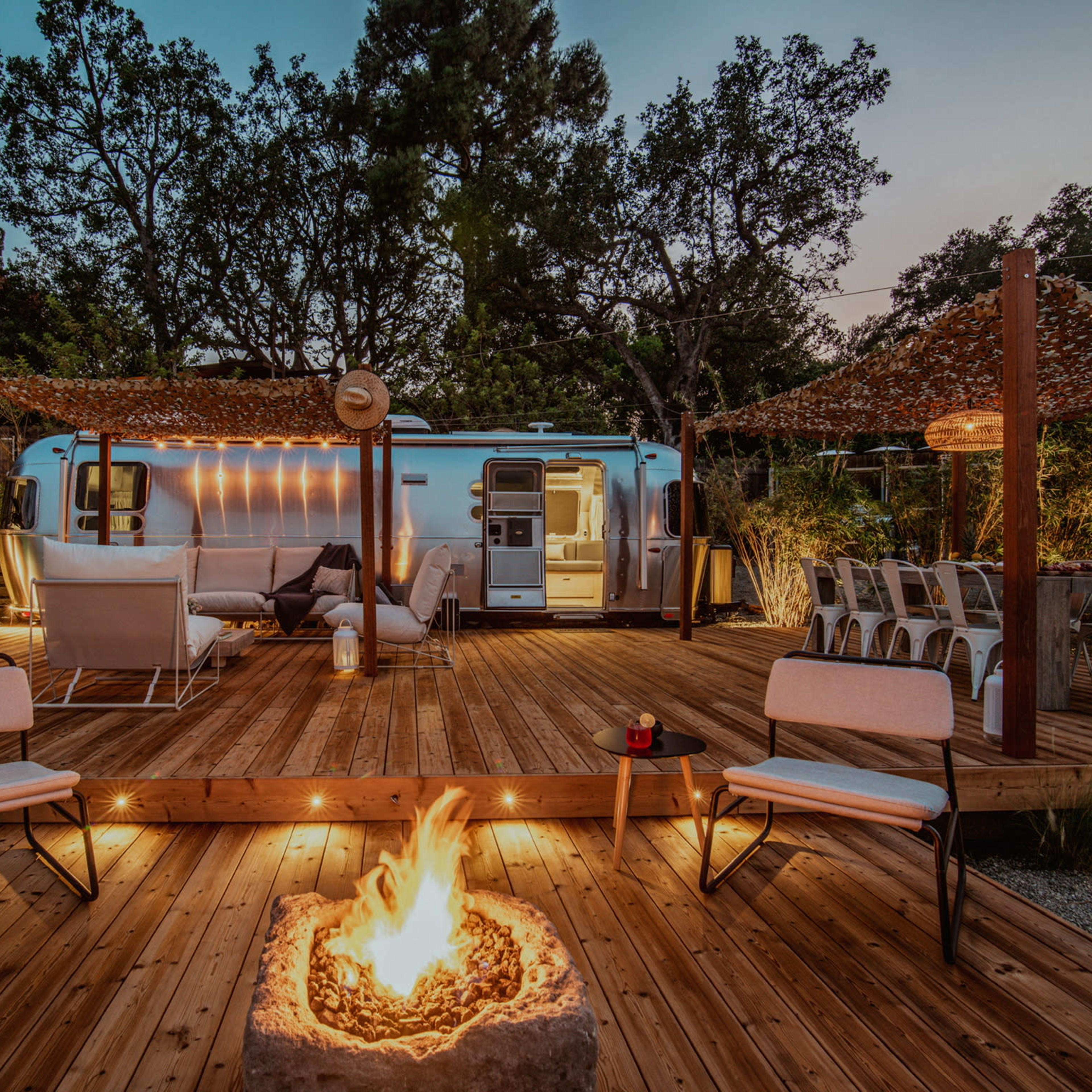 A cozy outdoor space features a fire pit surrounded by chairs, a deck with comfortable seating, and an Airstream trailer illuminated by string lights amidst trees.