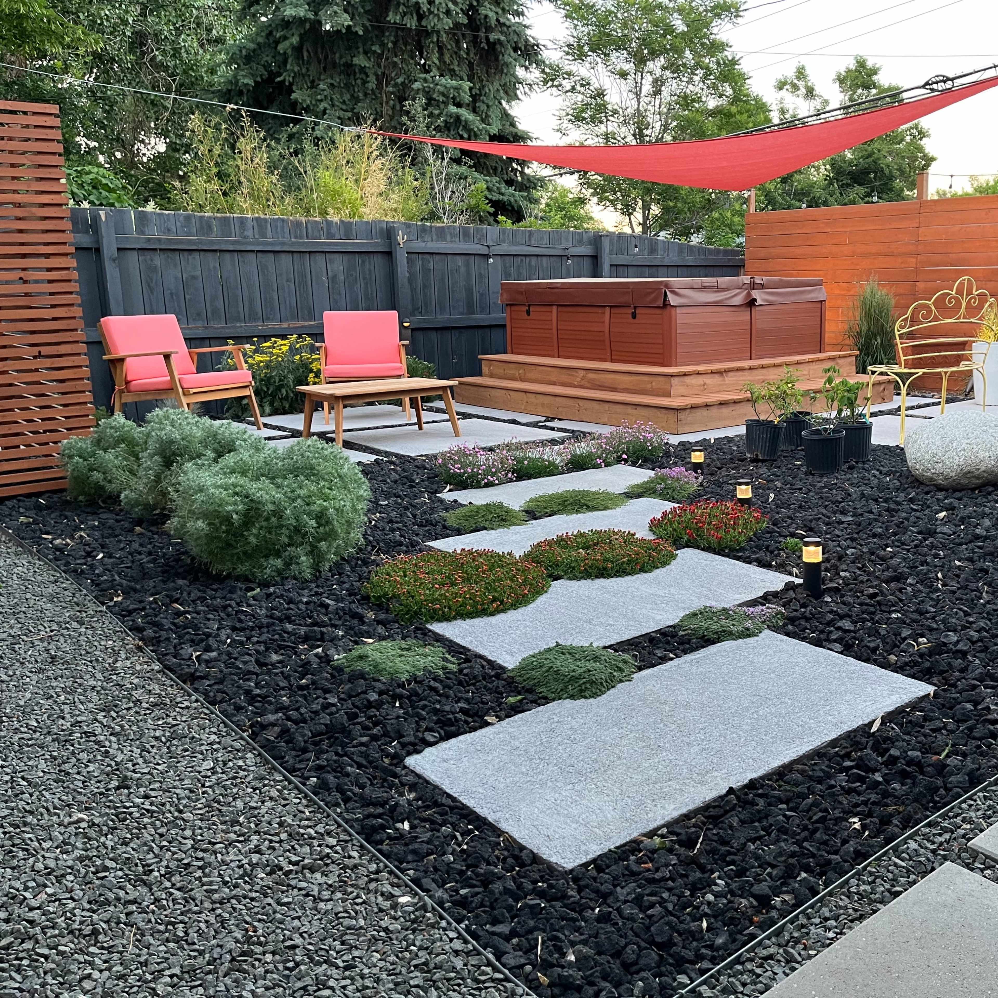 A landscaped backyard featuring a hot tub surrounded by stone pathways, decorative rocks, and two pink loungers under a red canopy.