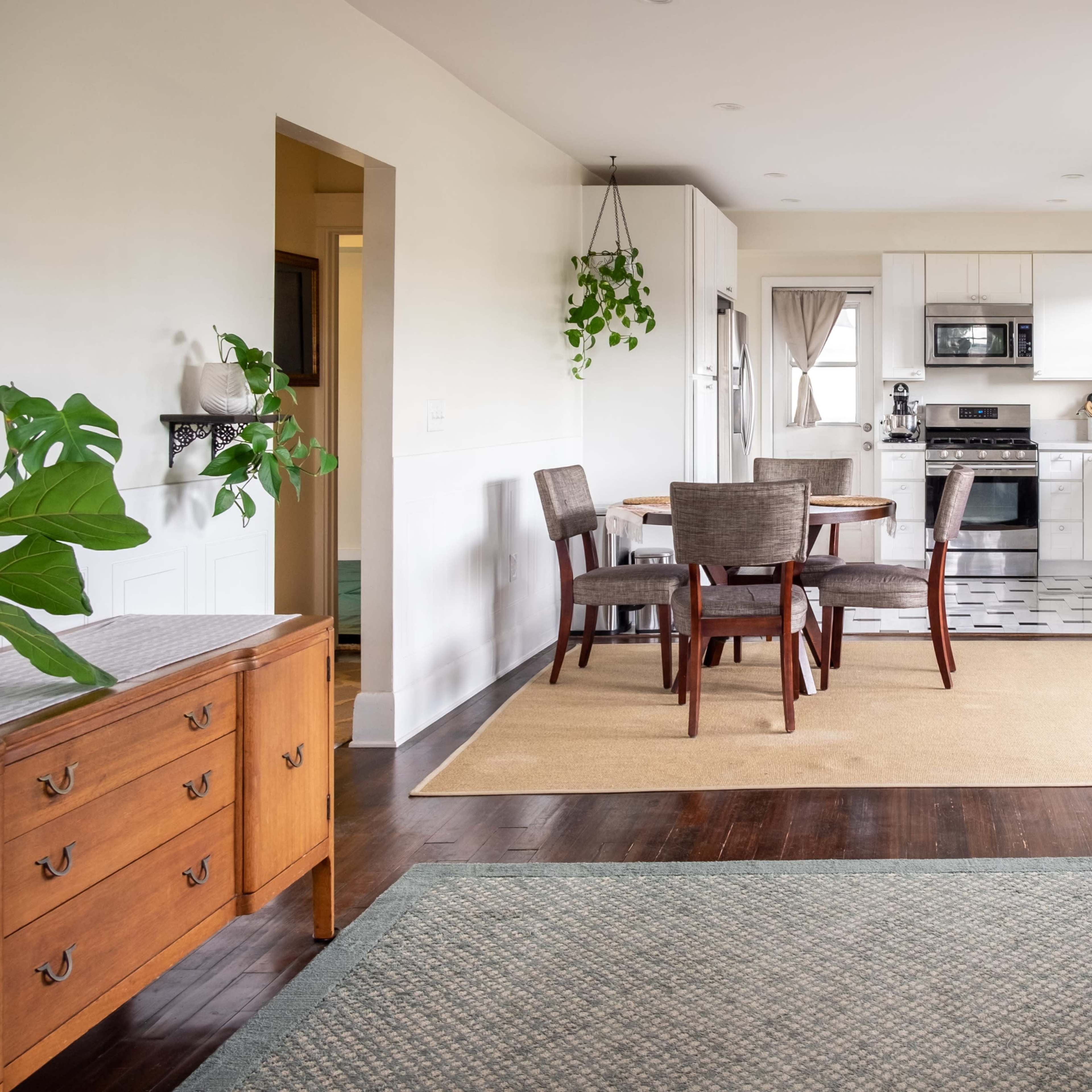 A modern, open-concept living and dining area features a wooden dining table with chairs, a sofa, and a kitchen visible in the background, framed by houseplants.