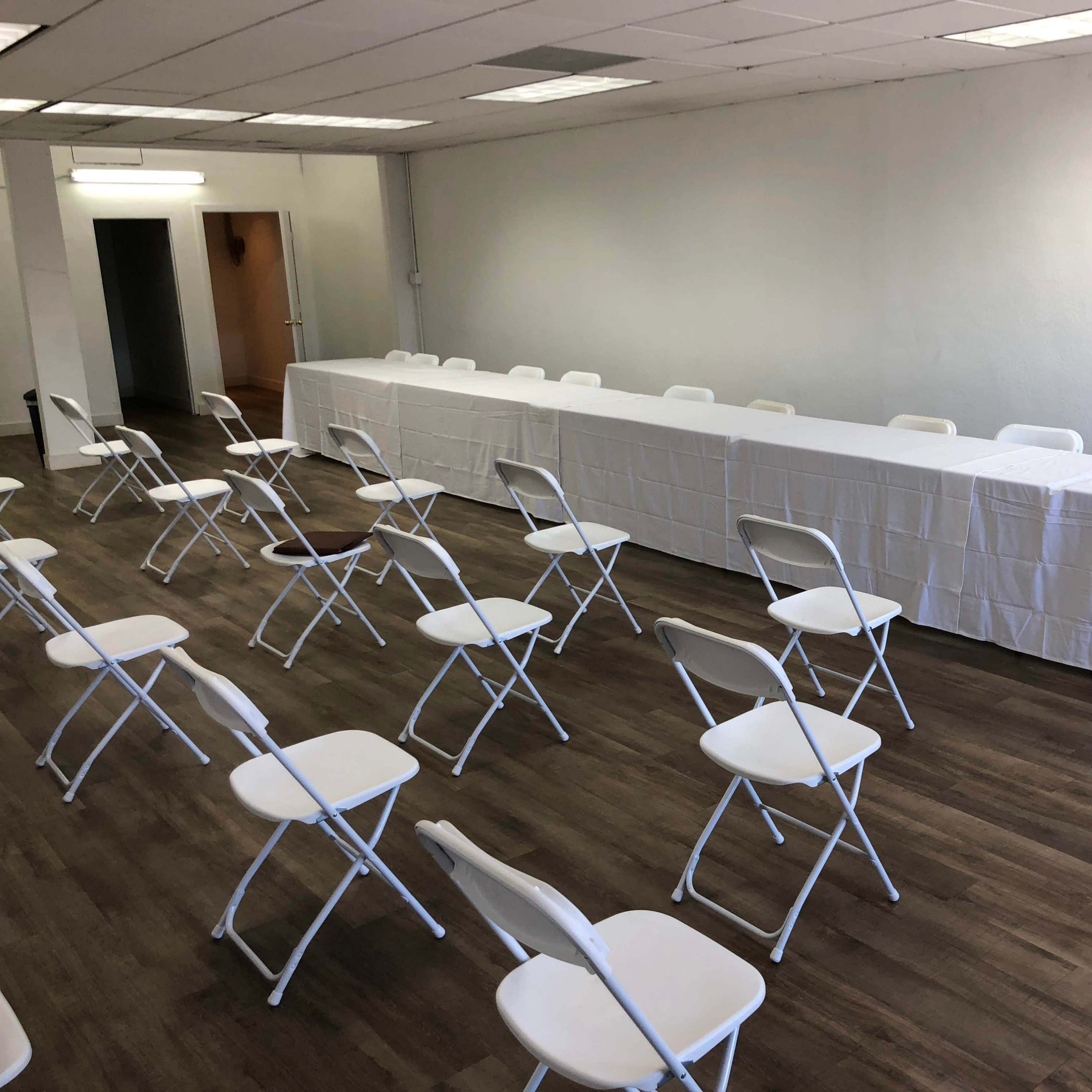 A large, empty room contains a long table covered with a white tablecloth and arranged folding chairs facing it.