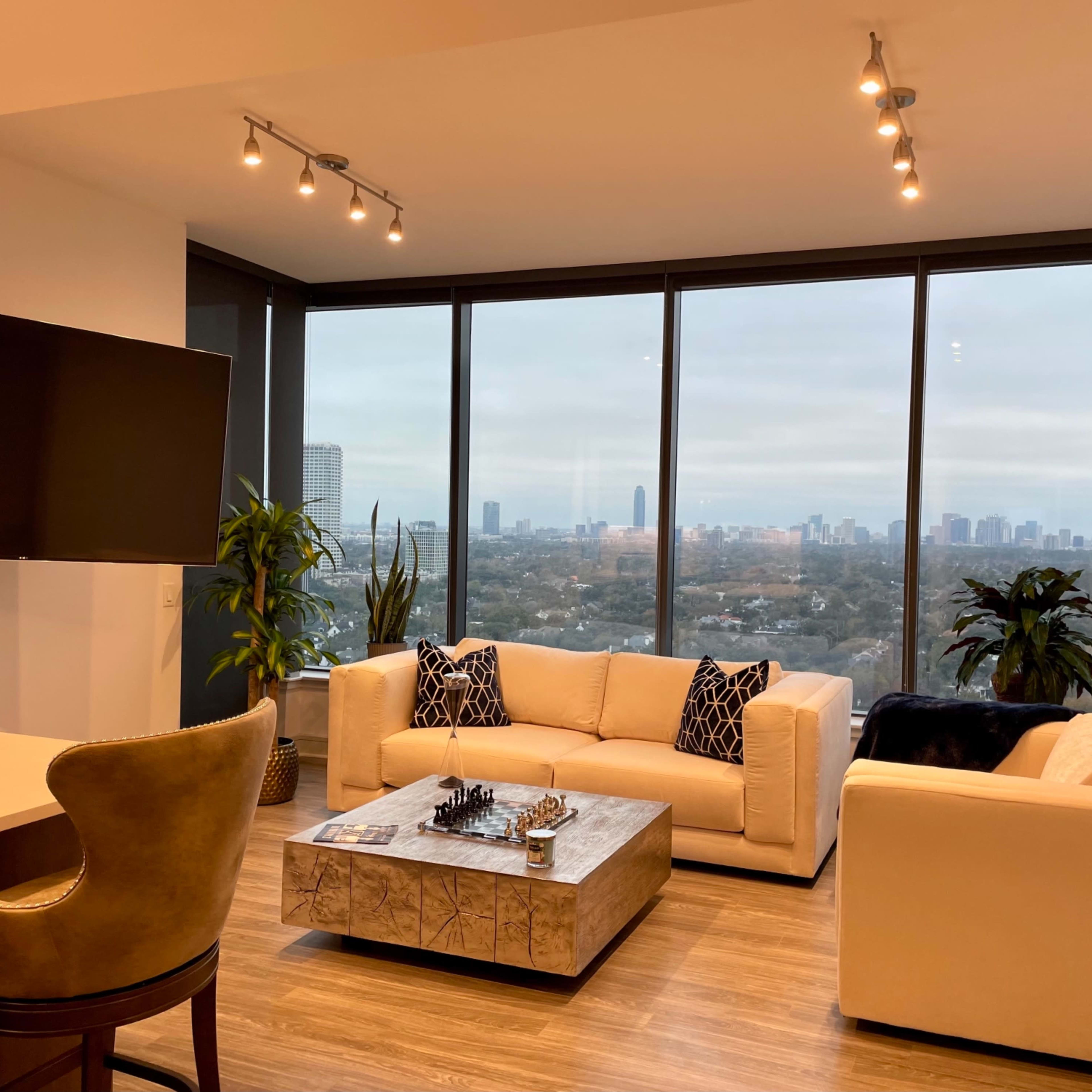 A modern living room features two sofas and a coffee table with a chess set, overlooking a city skyline through large windows.
