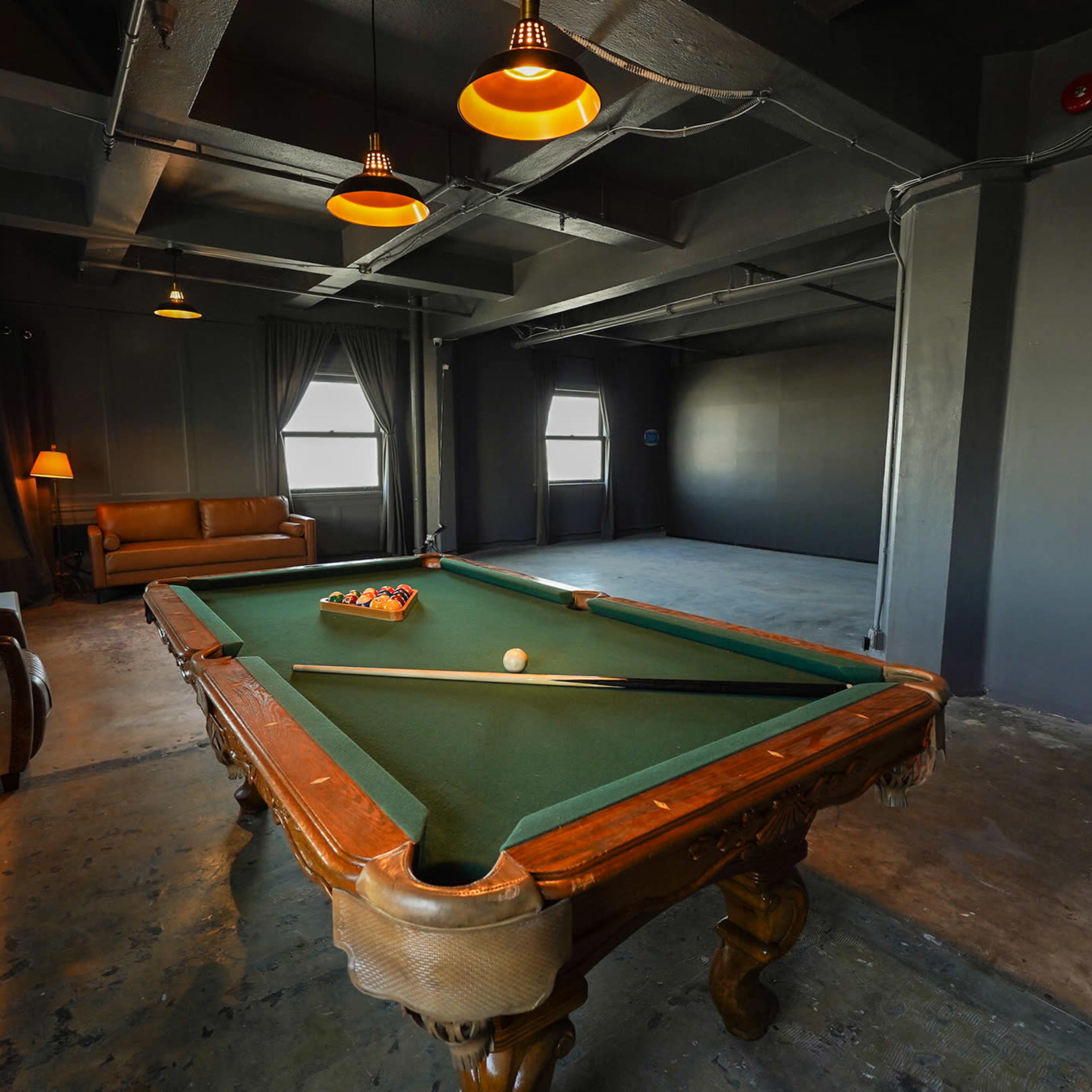A room with a billiards table set in the center, flanked by a sofa and an armchair, with windows and ceiling lights illuminating the space.