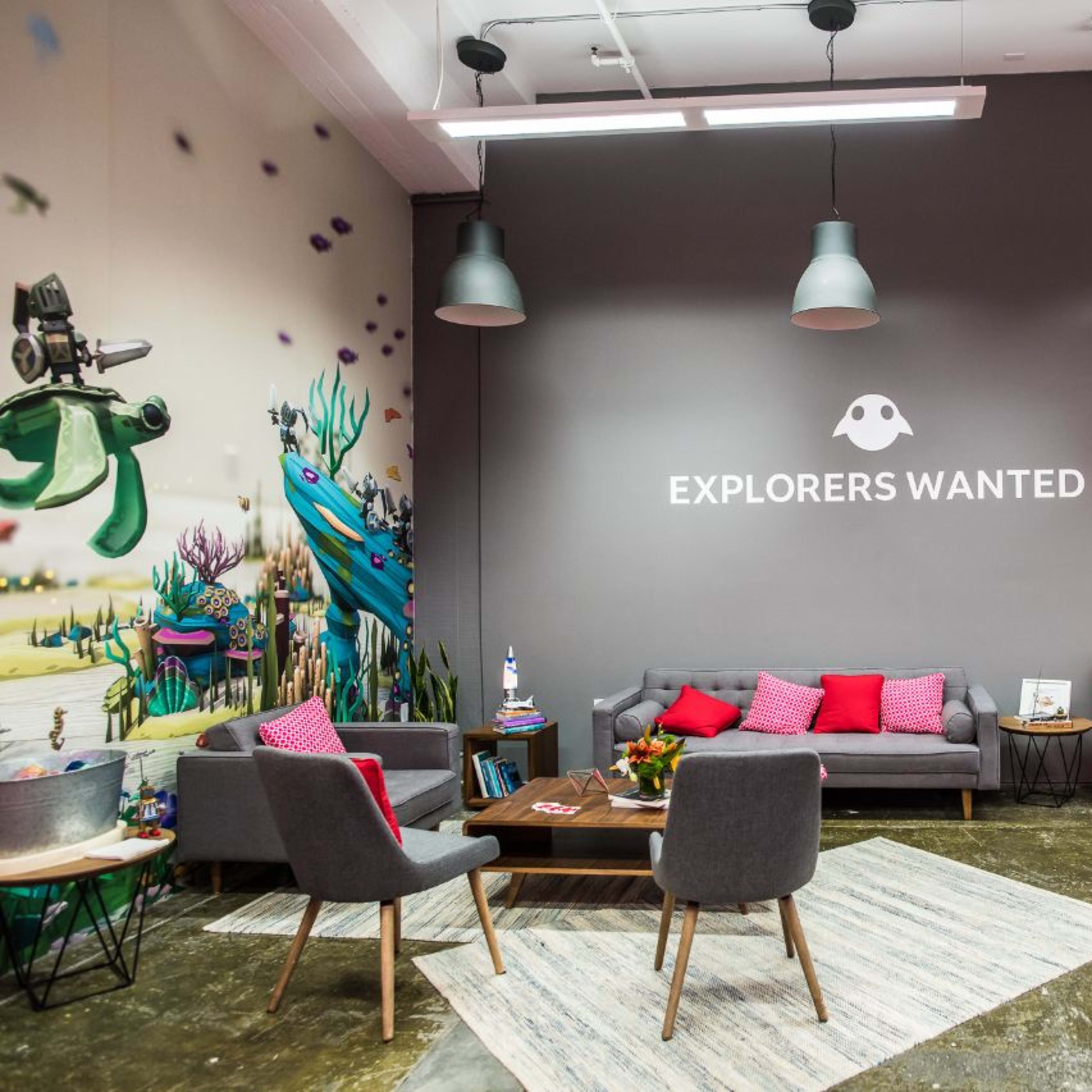 A modern lounge area featuring a gray wall with the phrase "EXPLORERS WANTED," a gray sofa with pink cushions, red chairs, and a decorative rug on a polished floor.