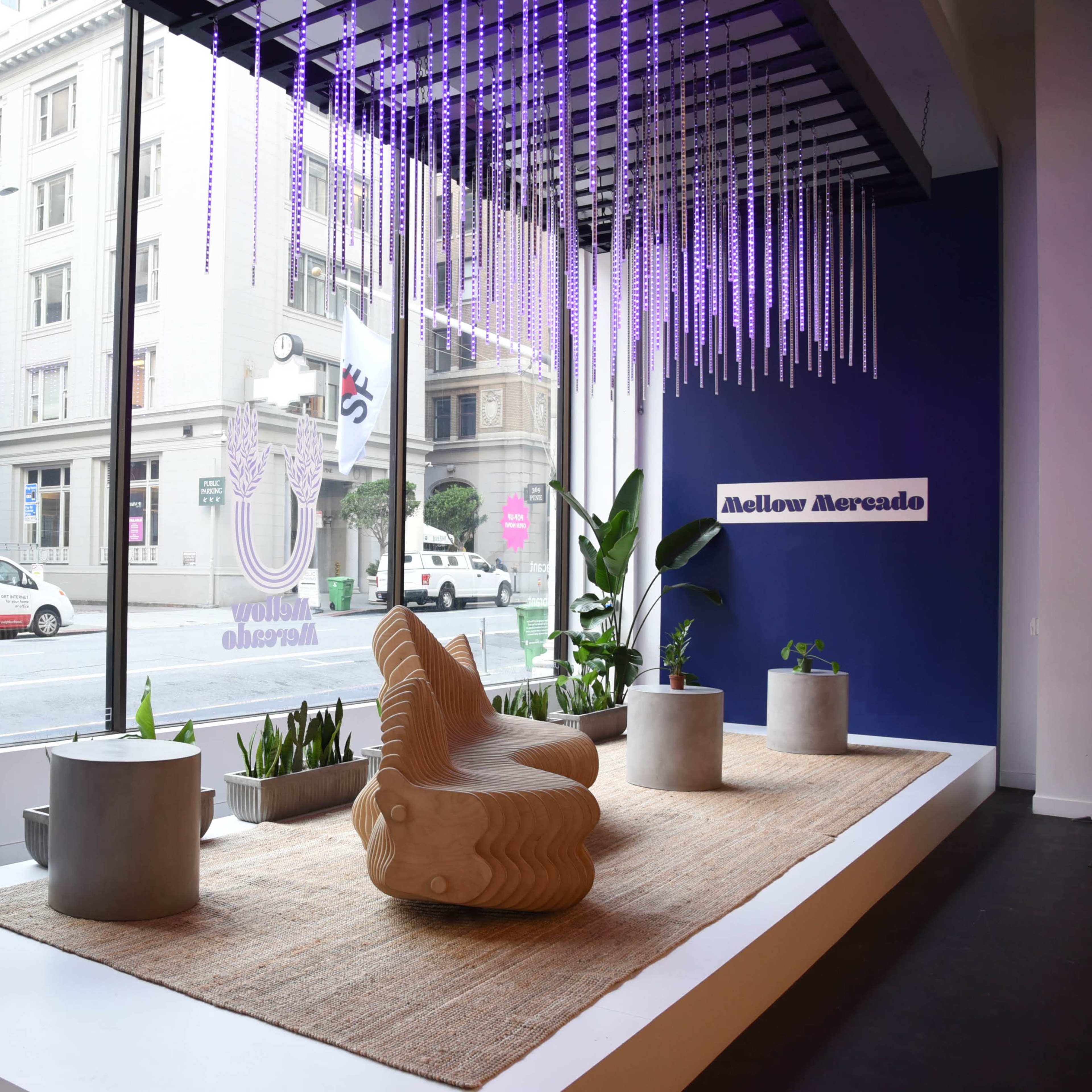 A modern interior space with a wooden chair, potted plants, and decorative lighting, situated by large windows overlooking a city street.