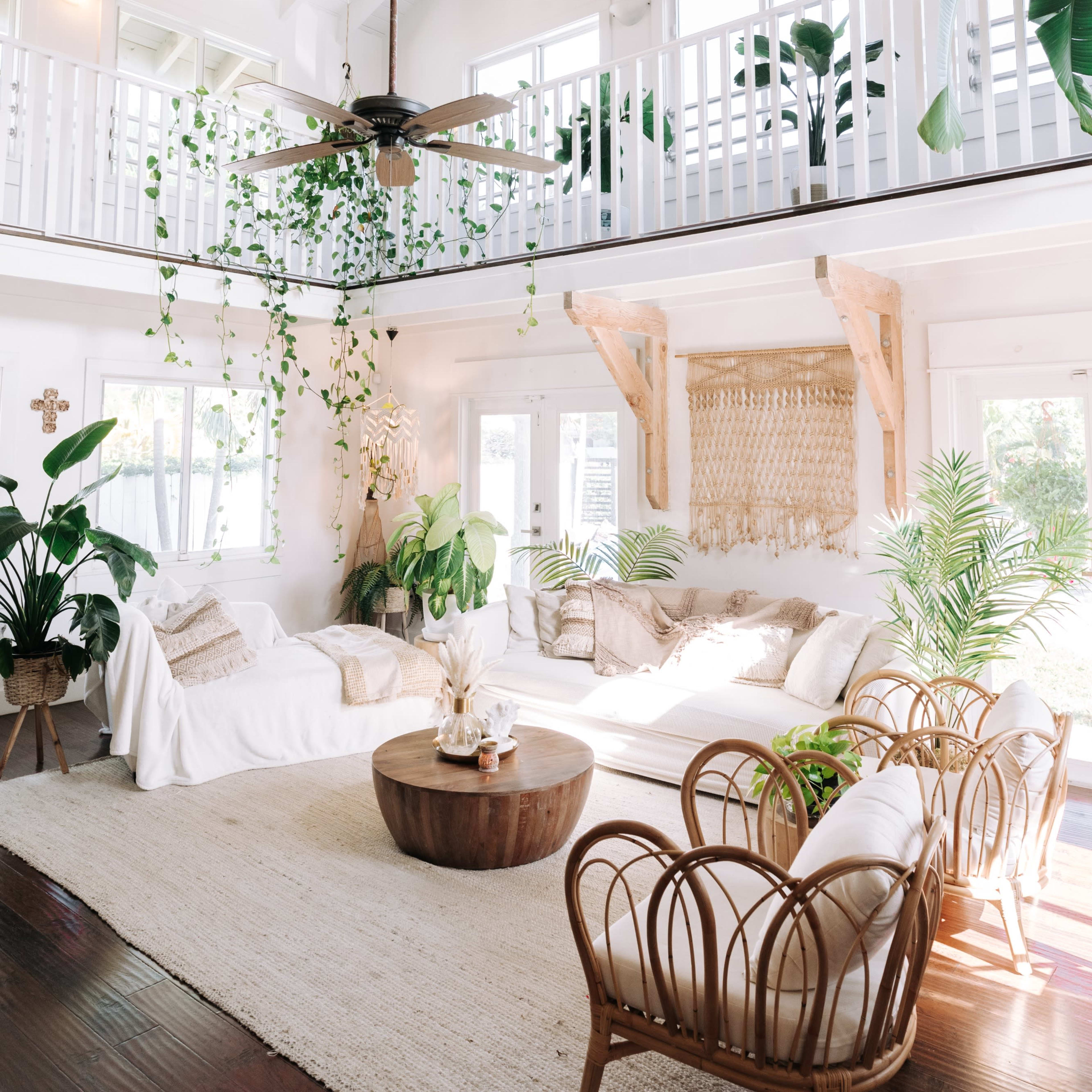 A spacious living room with high ceilings, large windows, and natural plants, featuring a circular wooden coffee table and woven chairs.