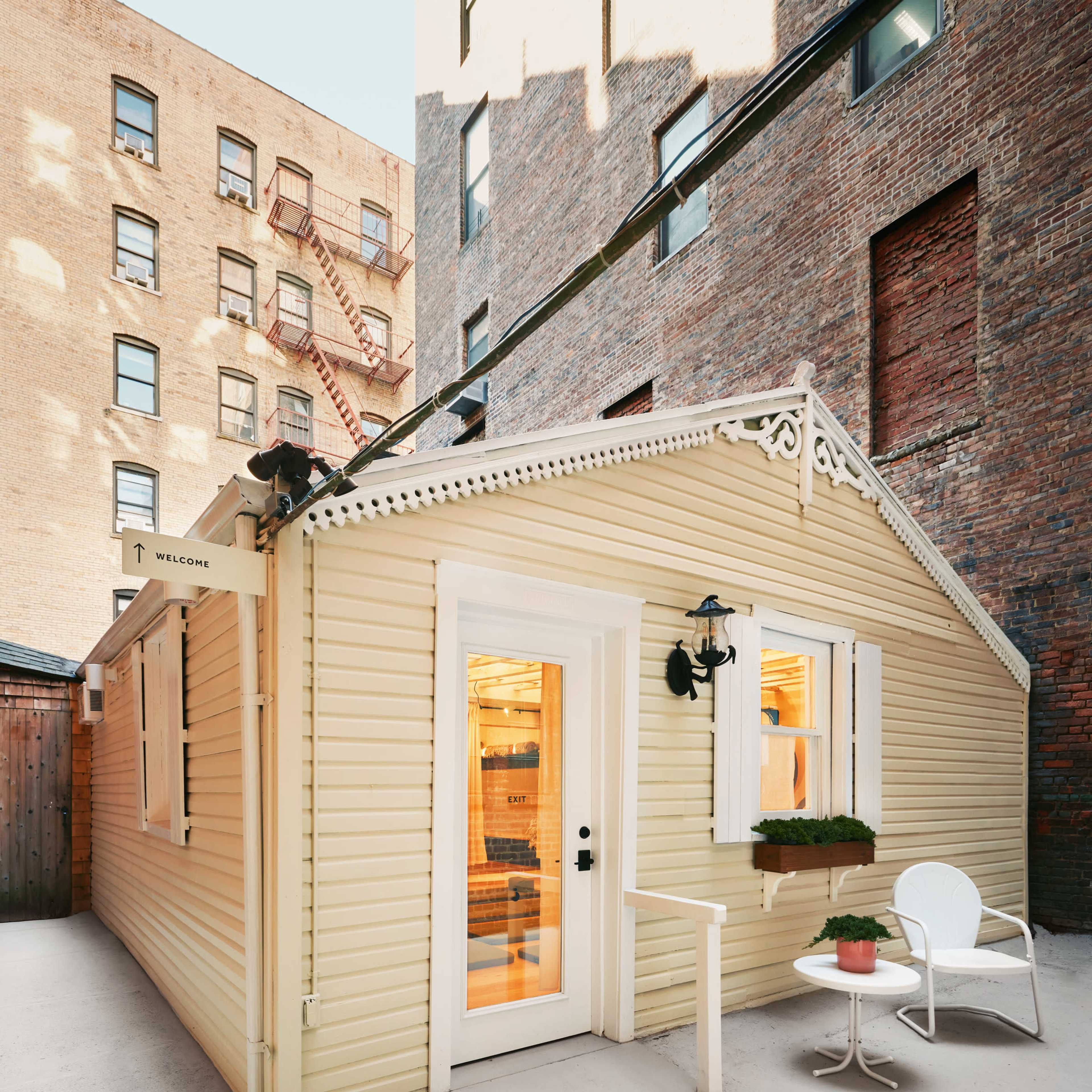 A small, yellow cottage with a white door and a chair set outside on a paved surface, surrounded by brick buildings.