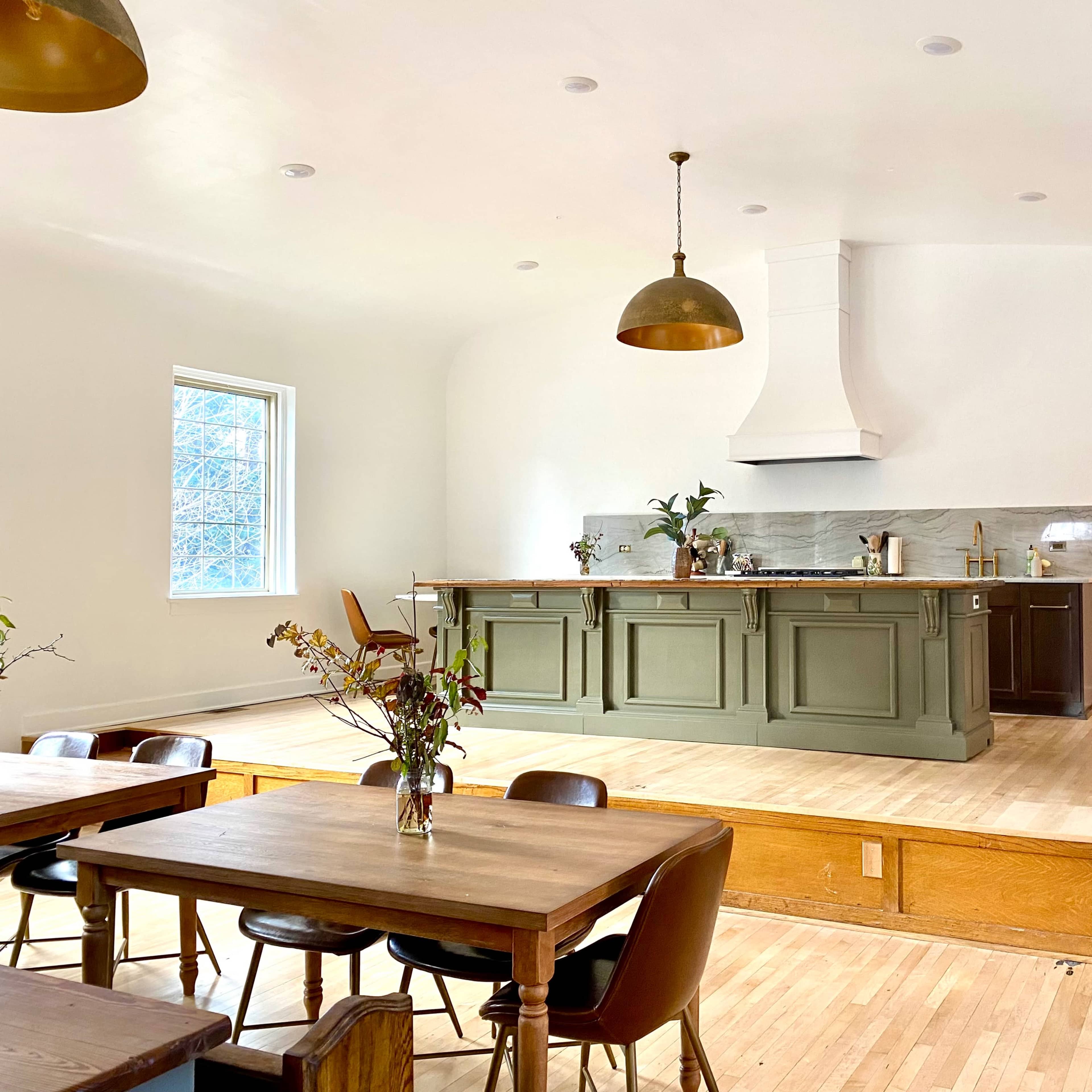A spacious kitchen features a green island with bar seating, a large hood above the stove, and wooden tables set for dining.