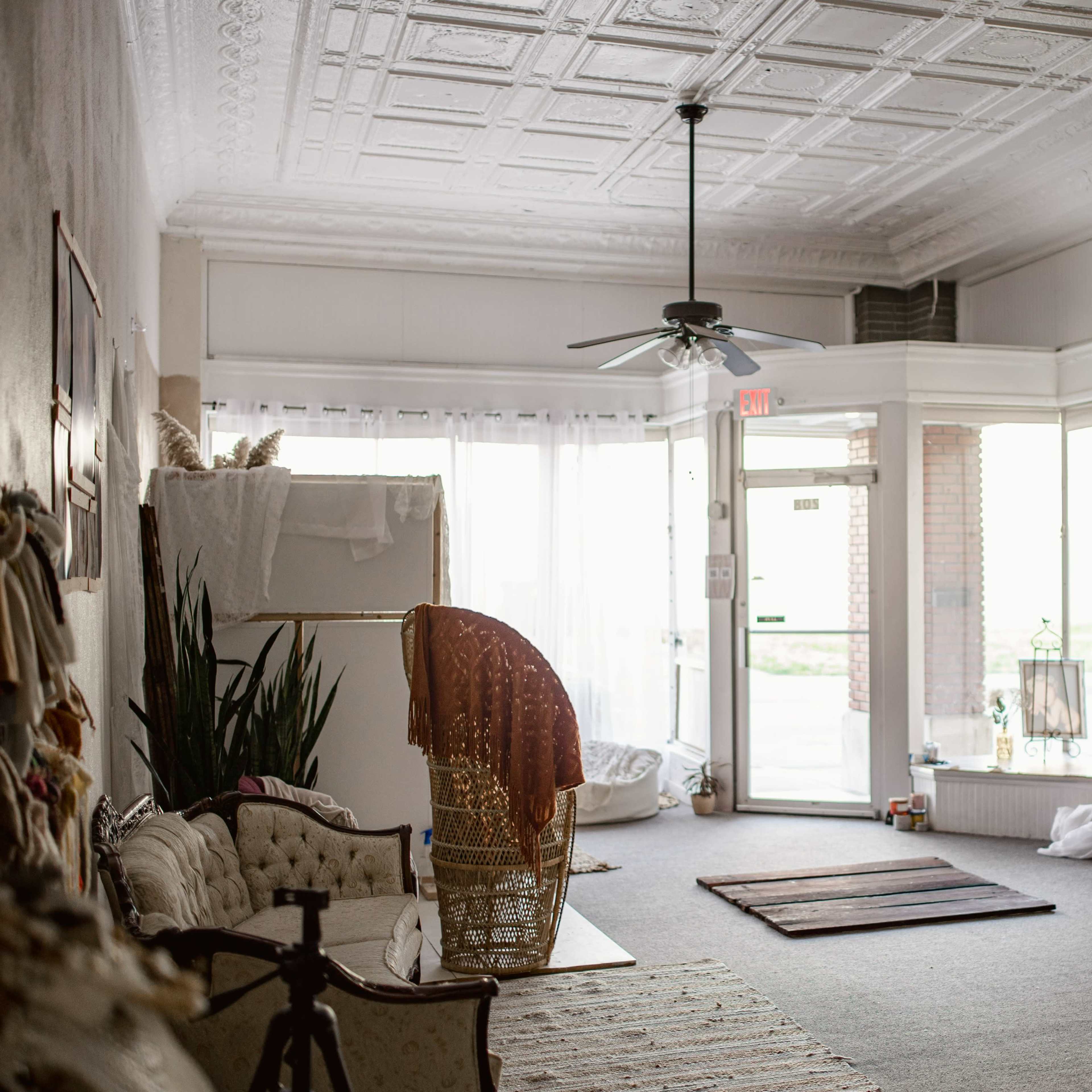 A spacious and empty room with a textured ceiling, featuring a seating area, plants, and large windows letting in natural light.