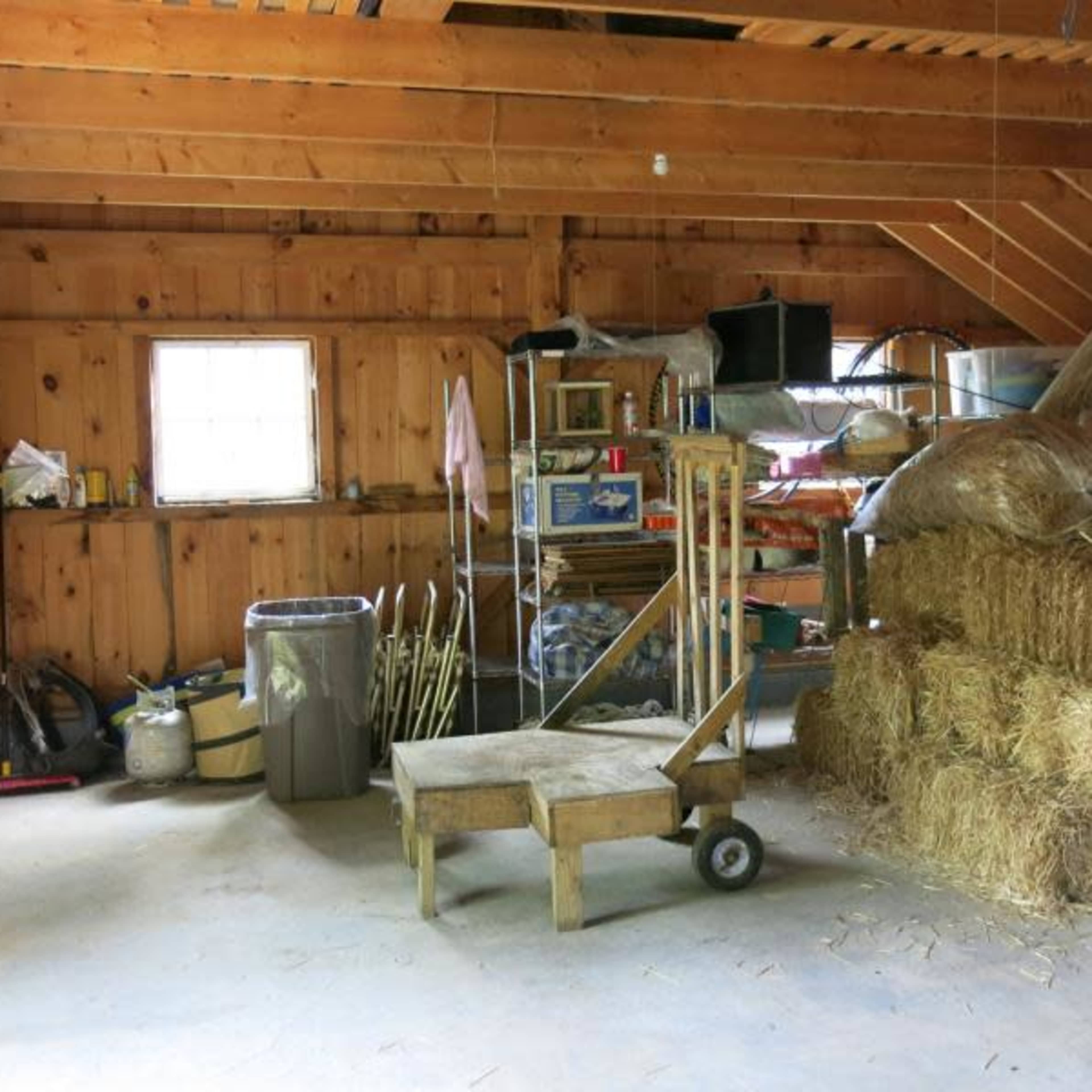 The interior of a wooden barn features hay bales stacked in one corner, shelves filled with tools, and a cart in the middle of the floor.