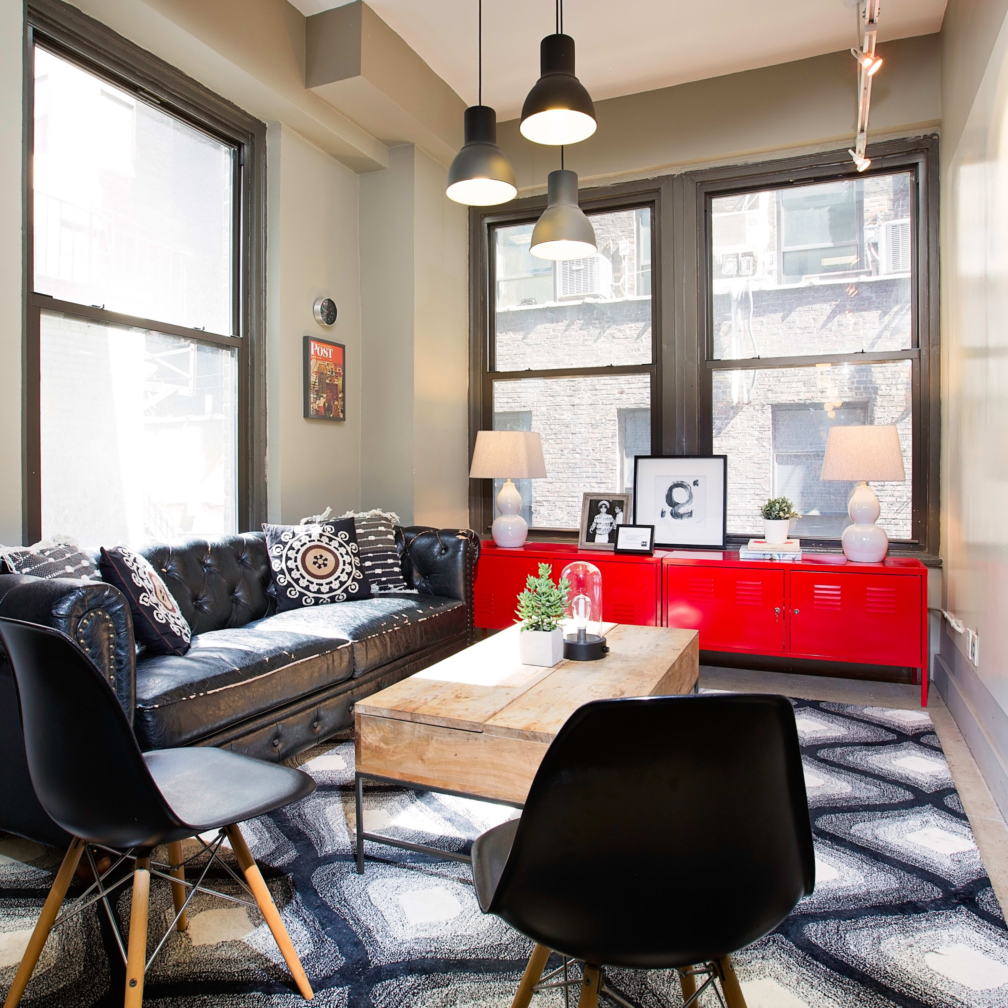 A modern living room with a black leather sofa, a wooden coffee table, and two chairs, featuring large windows that let in natural light.