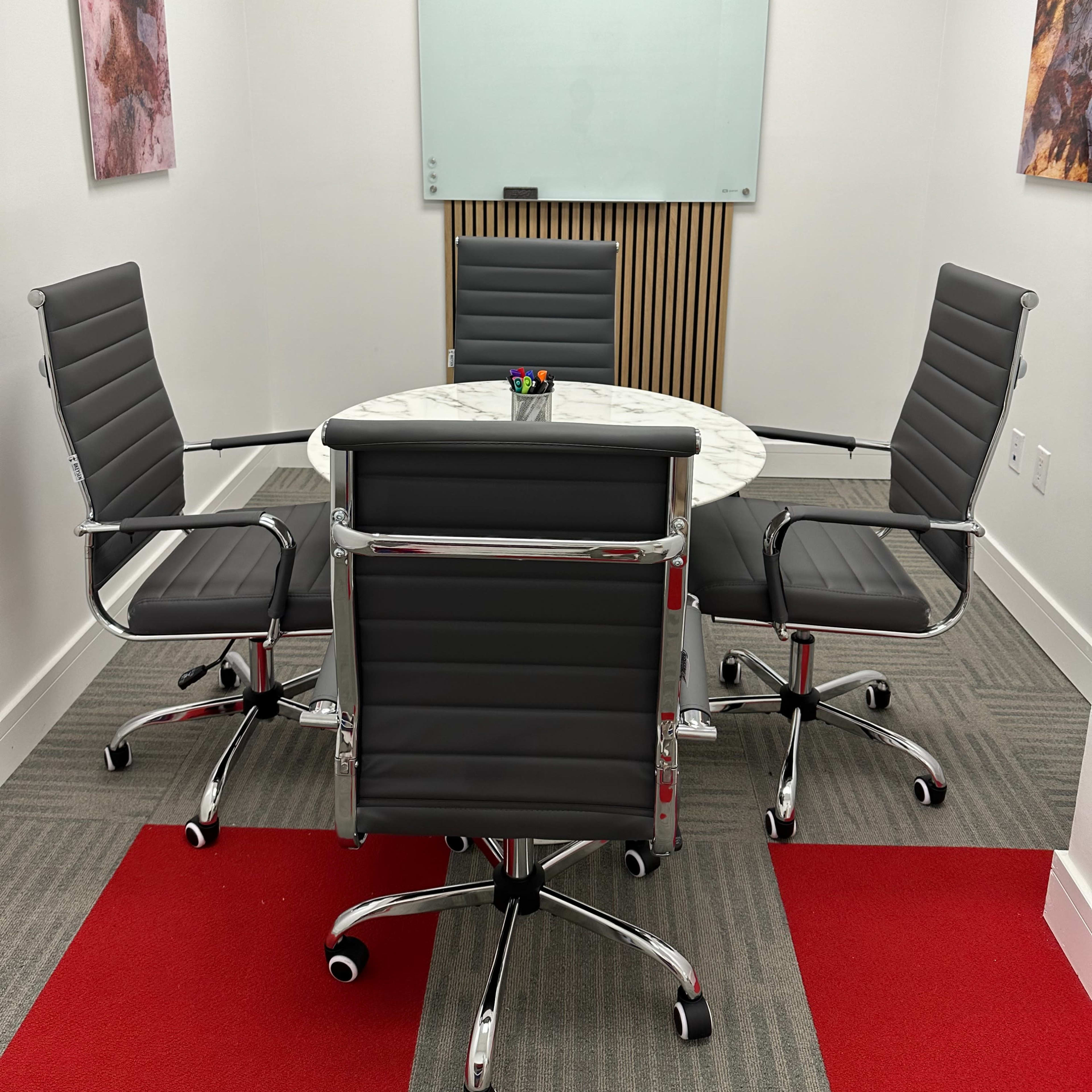A small conference room with a round marble table and four black leather chairs, flanked by white walls and wooden paneling.