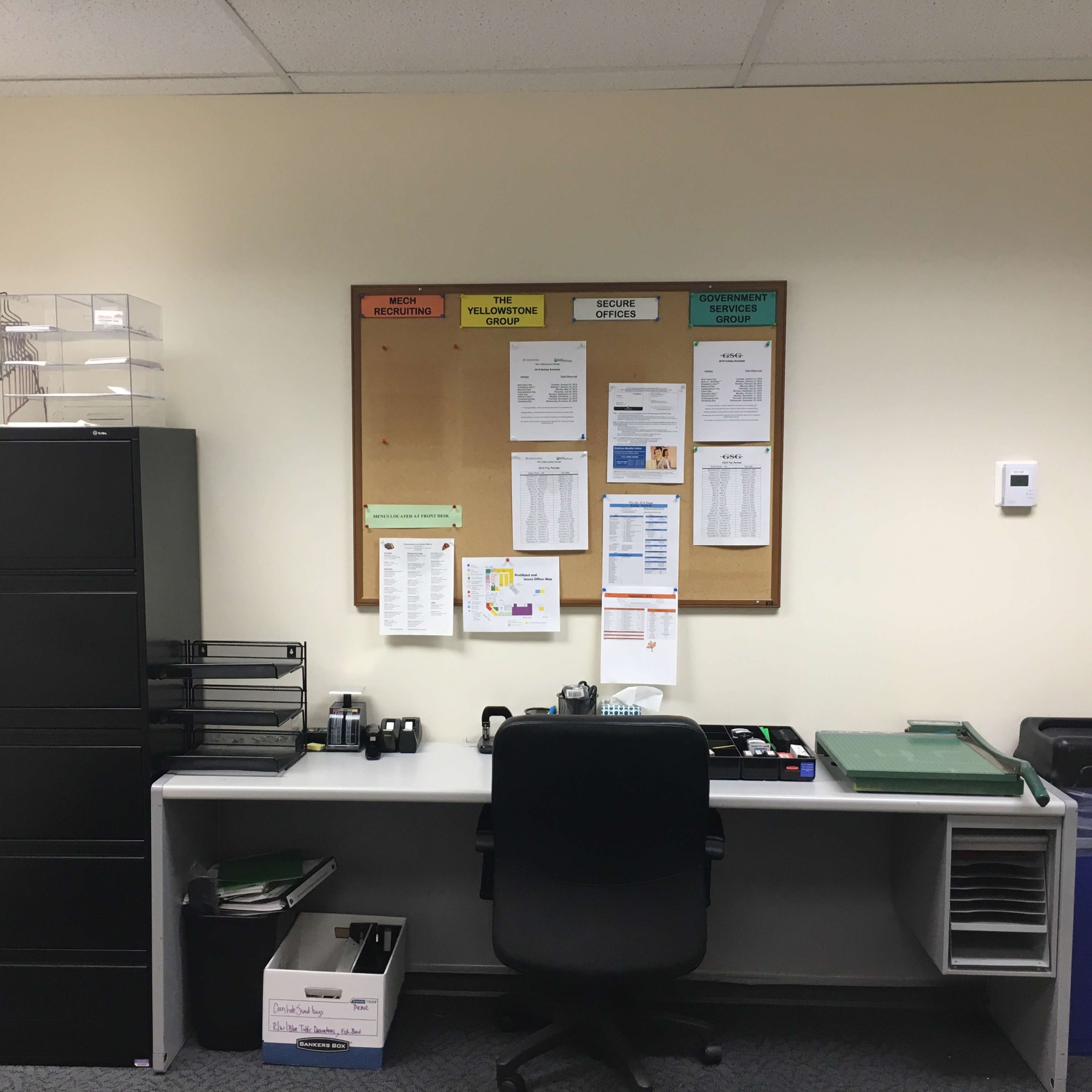 A small office with a desk, an office chair, a filing cabinet, and a bulletin board displaying various documents and notices.