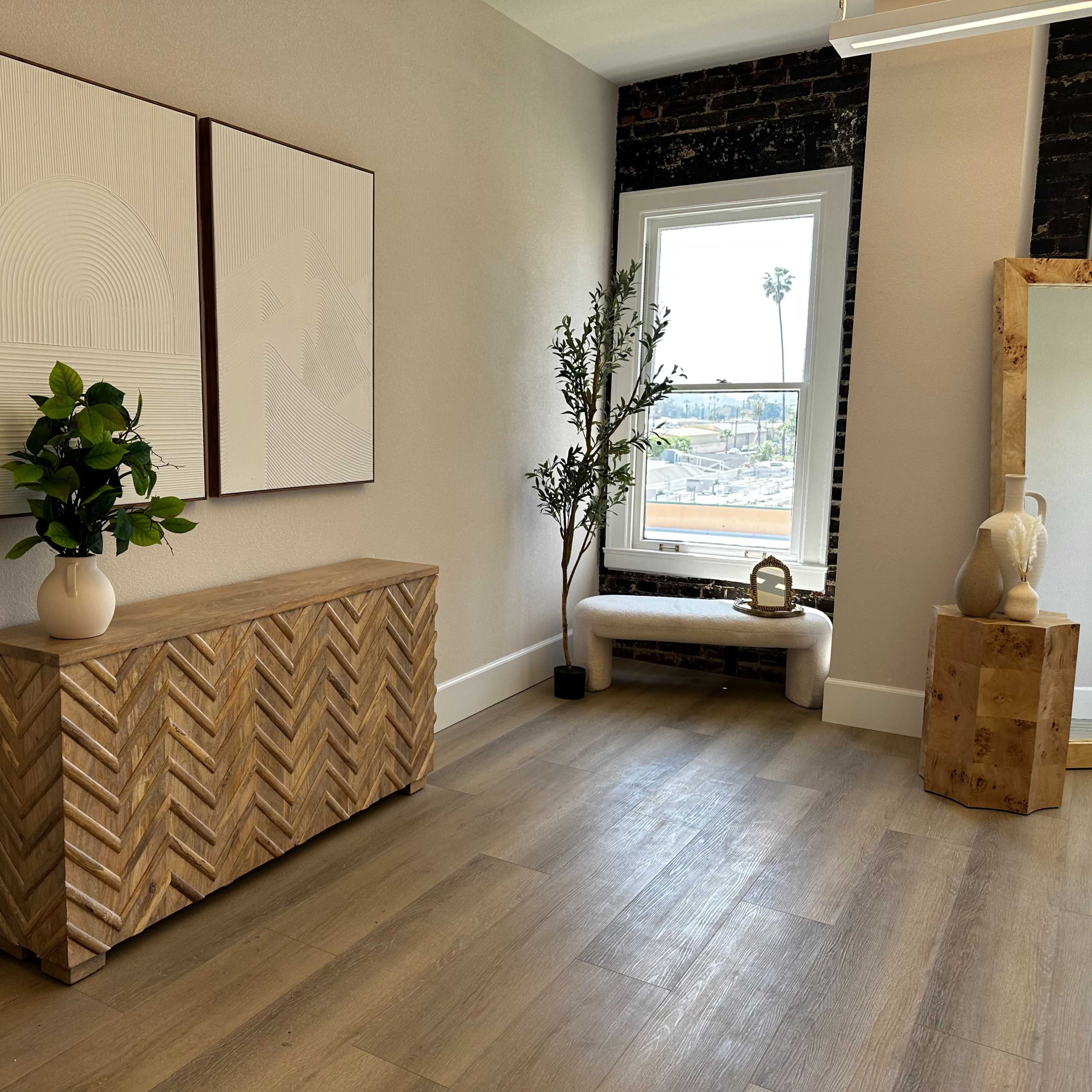 A room with a wooden cabinet featuring a herringbone pattern, two framed artworks on the wall, a potted plant, and large windows with a view of palm trees outside.