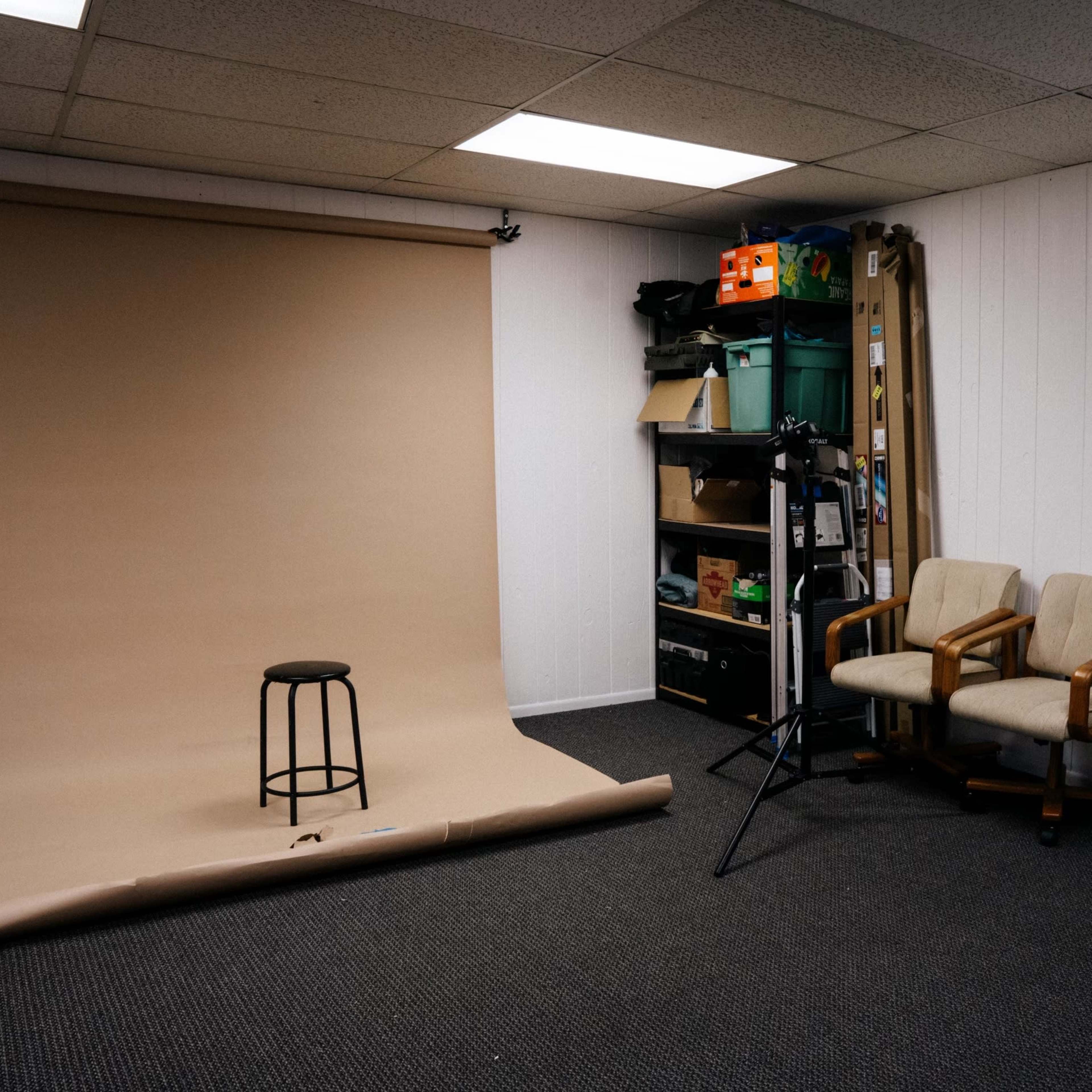 A photography studio with a beige backdrop, a black stool in the center, two chairs against the wall, and storage shelves filled with various items.