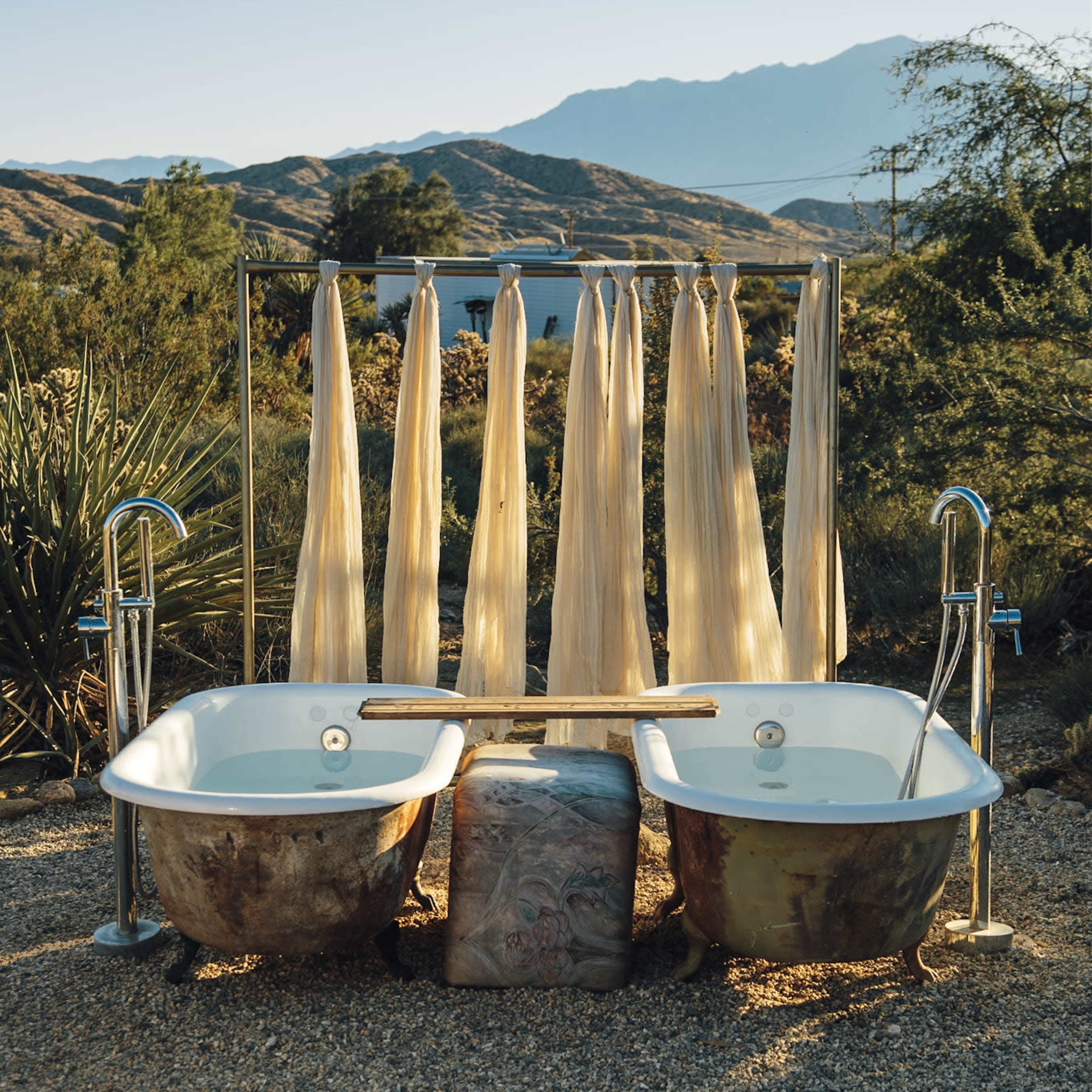 Two vintage bathtubs with a wooden plank between them sit in a gravel area, flanked by long fabric curtains against a backdrop of mountains and greenery.