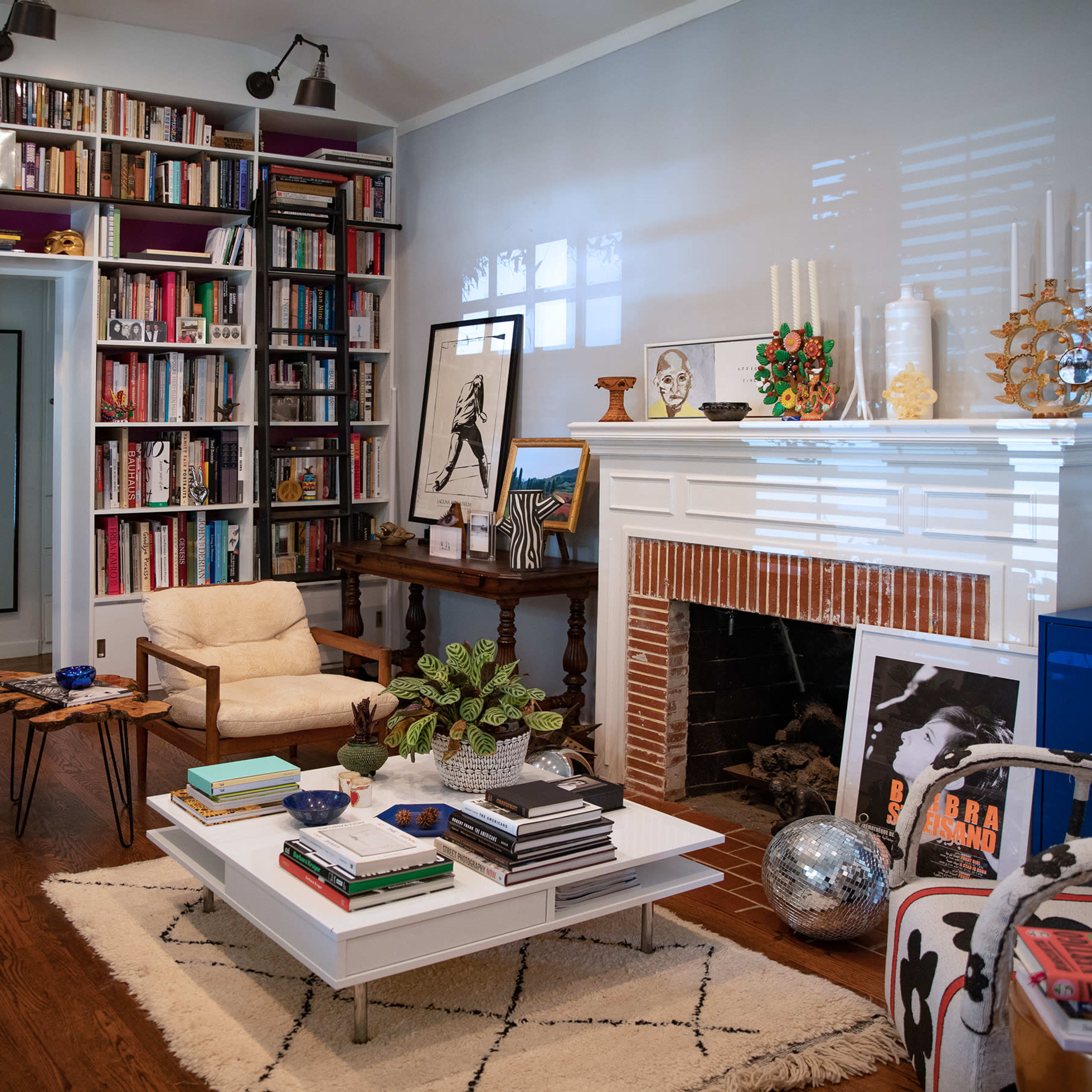 A cozy living room featuring a large bookshelf filled with books, a cream-colored armchair, a coffee table with various items, and a fireplace decorated with artwork.