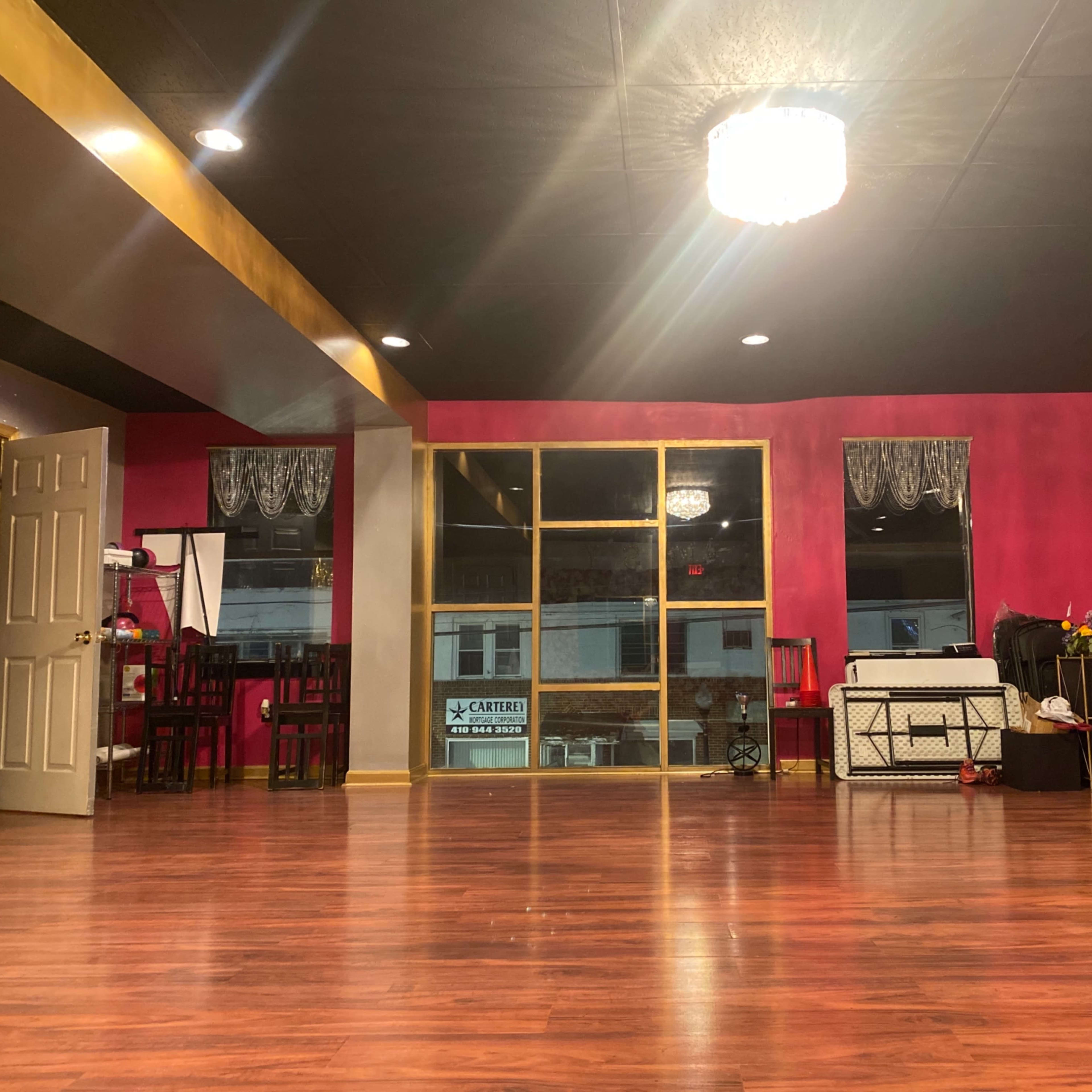 A spacious dance studio with a polished wooden floor and pink walls, featuring large windows and various pieces of furniture along the edges.