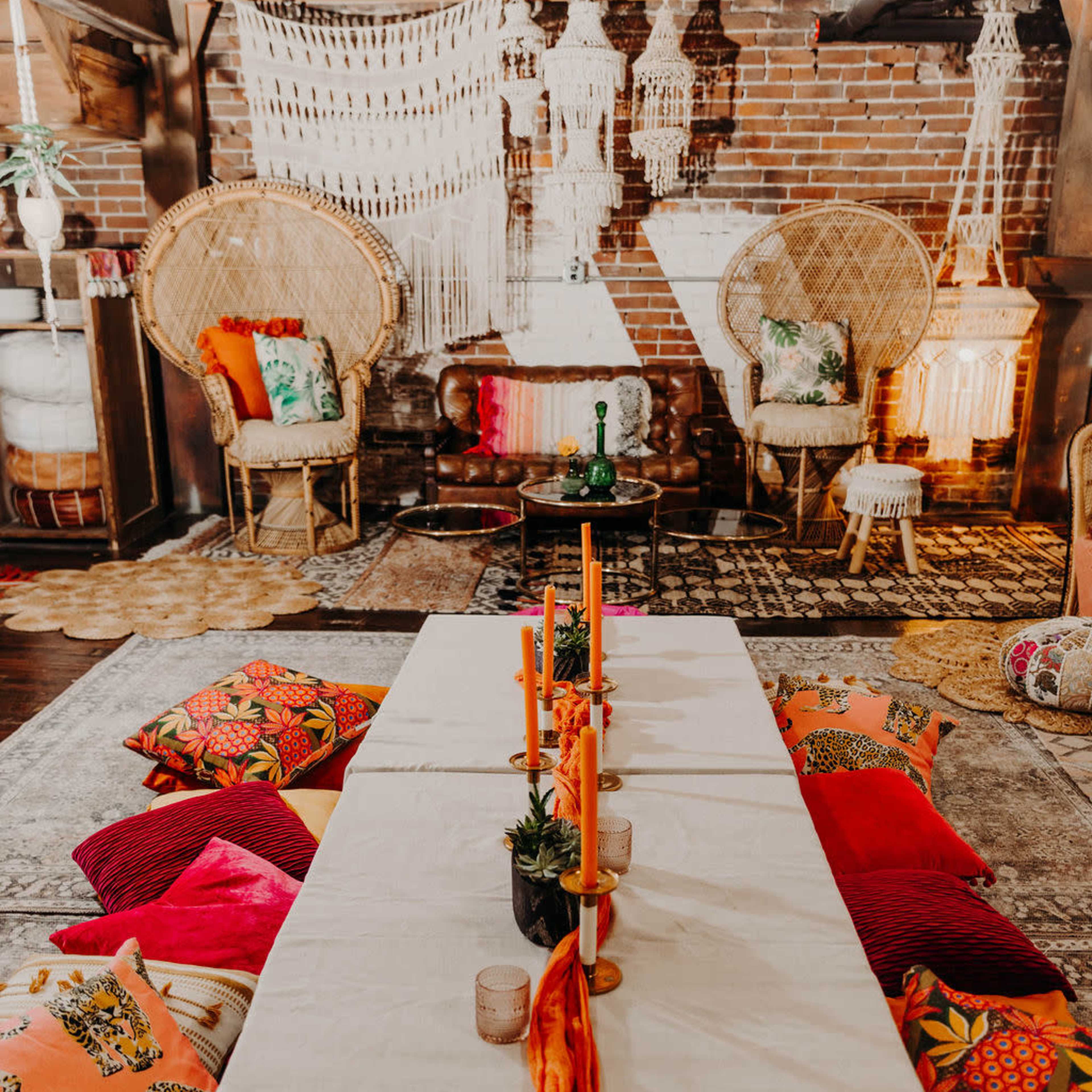 A decorated indoor space featuring a long table set with candles and surrounded by vibrant cushions and two large, woven chairs.