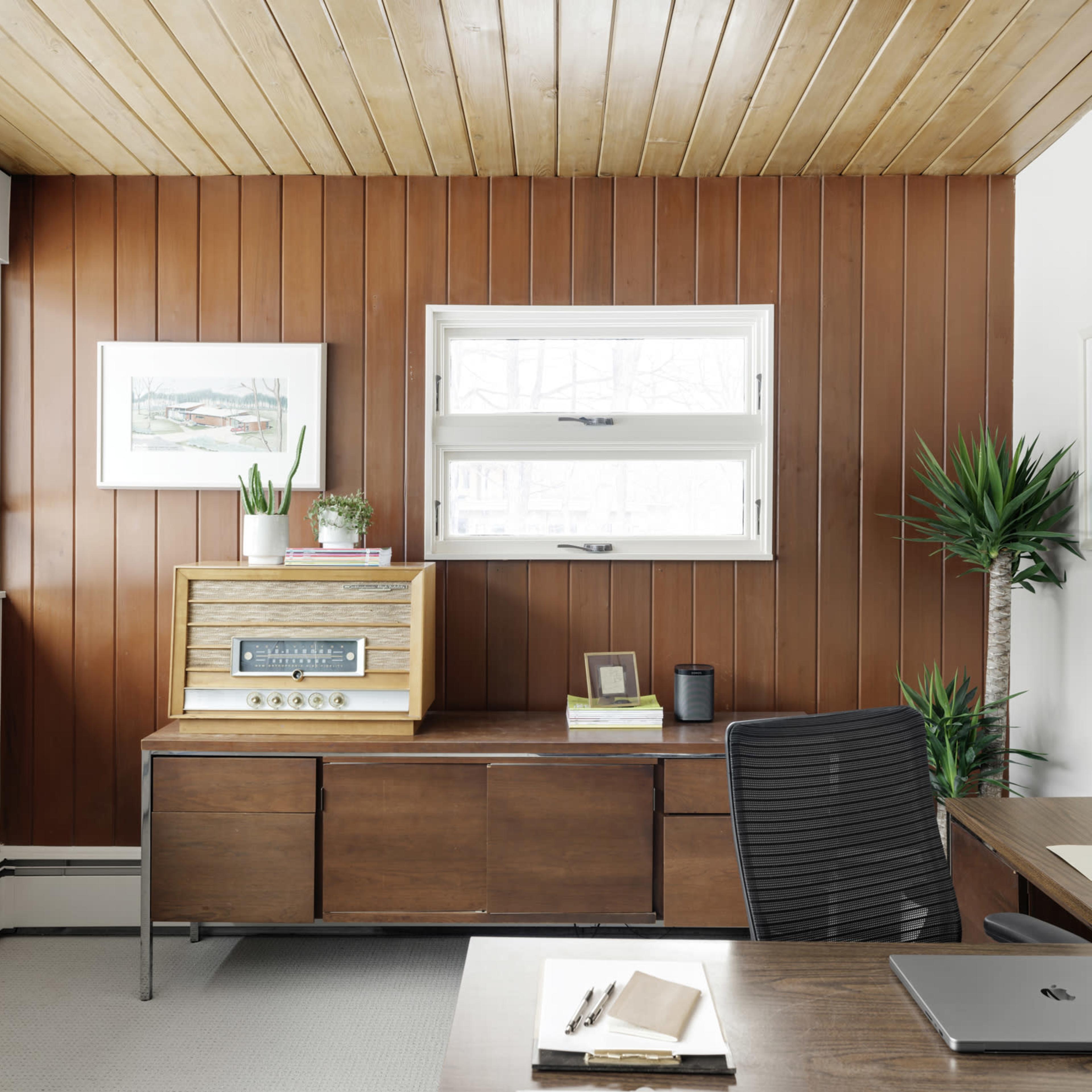 A modern office features wooden paneling, a desk with a laptop, a retro-style record player, and plants near the windows.