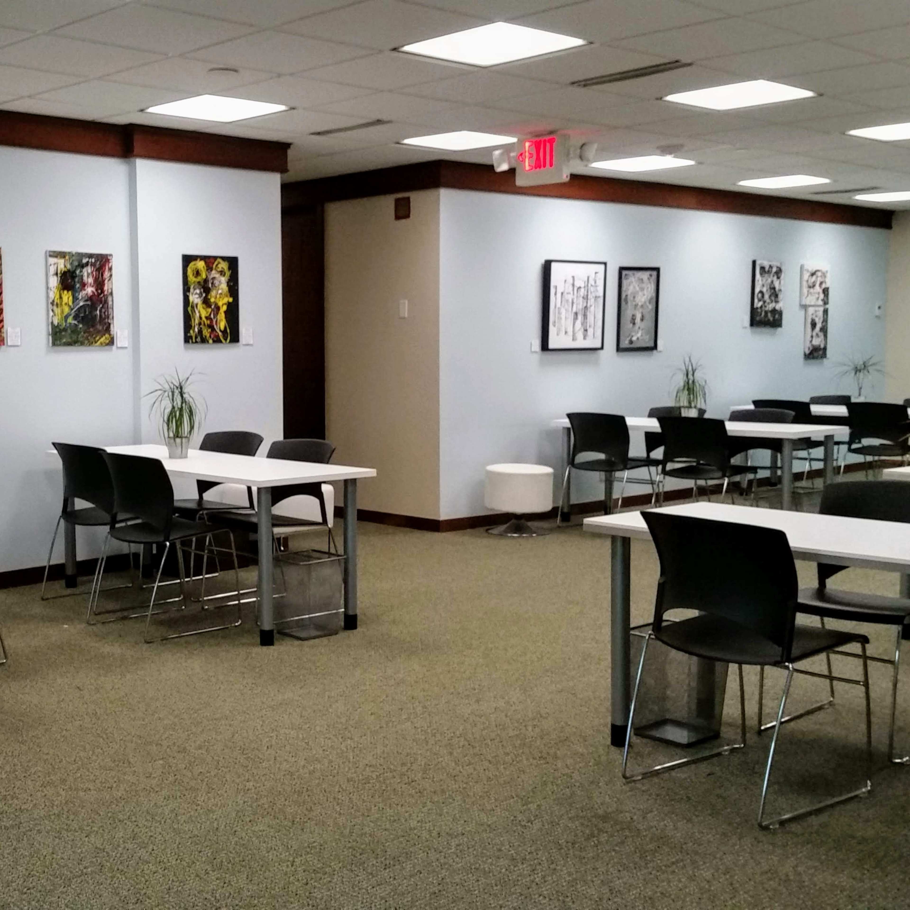 A spacious room with several tables and chairs, featuring artwork on the walls and plants on the tables.