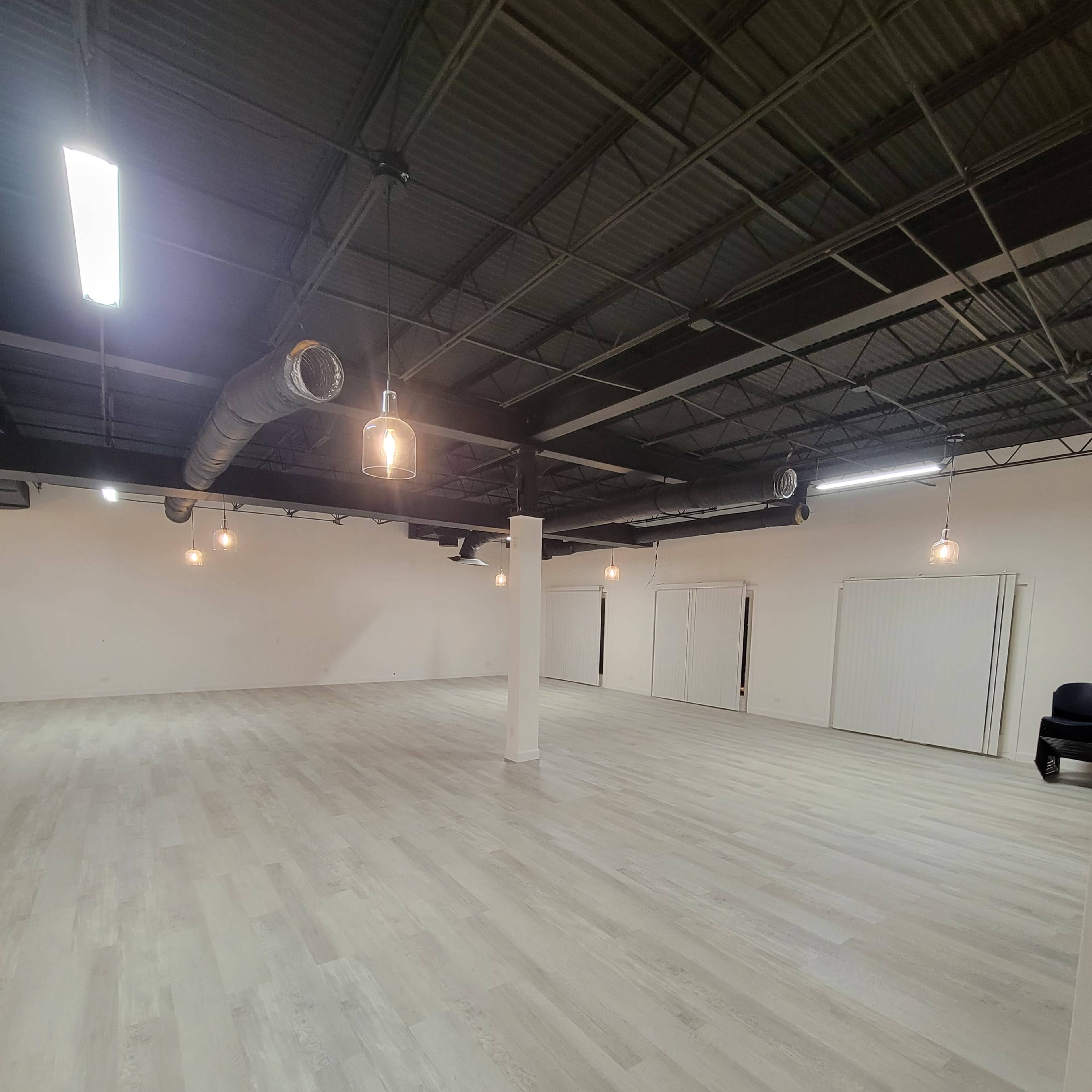 A spacious empty room with light-colored flooring, exposed ductwork, and overhead lighting fixtures.