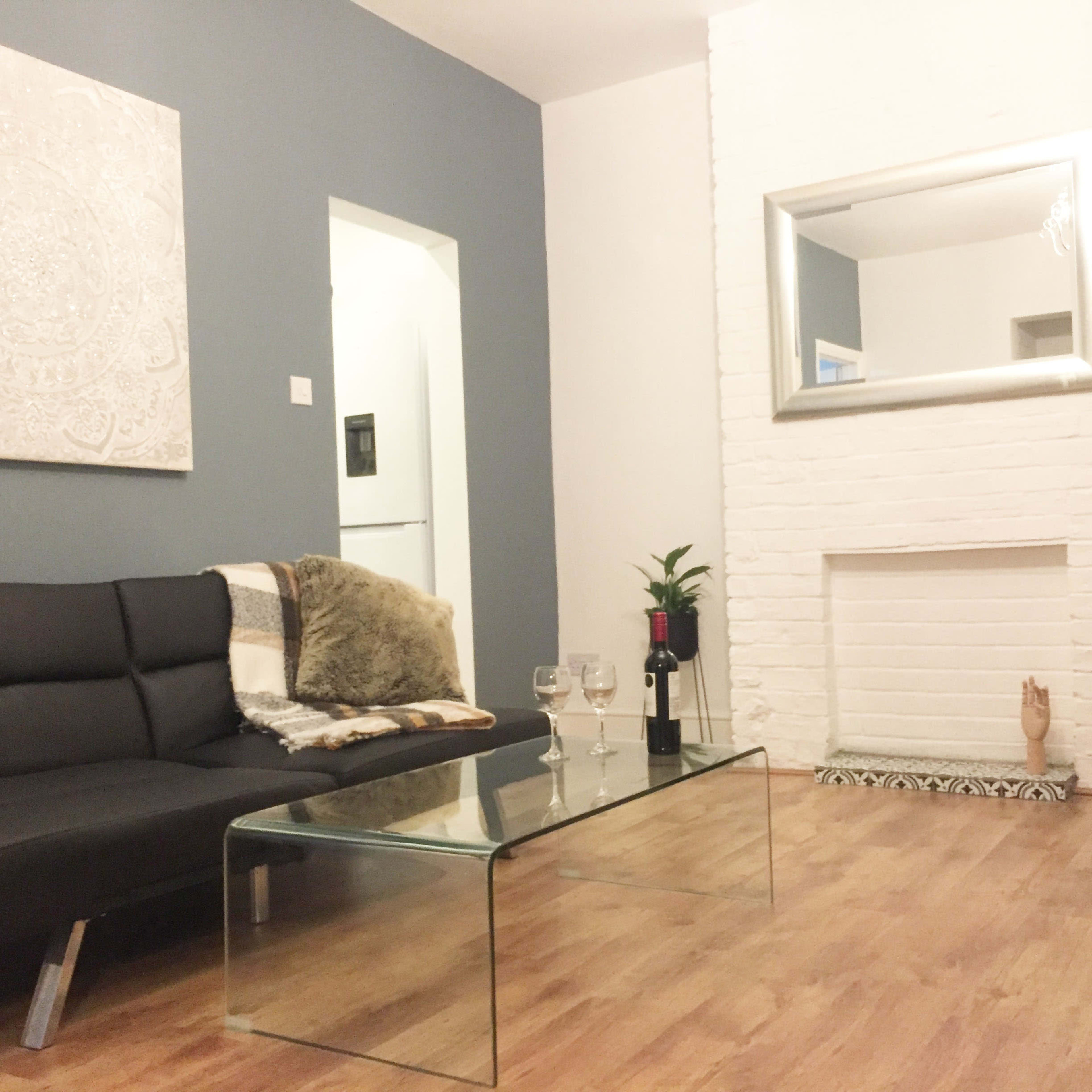 A modern living room featuring a black sofa, a glass coffee table with two wine glasses, and a decorative plant near a white fireplace with a mirror above it.