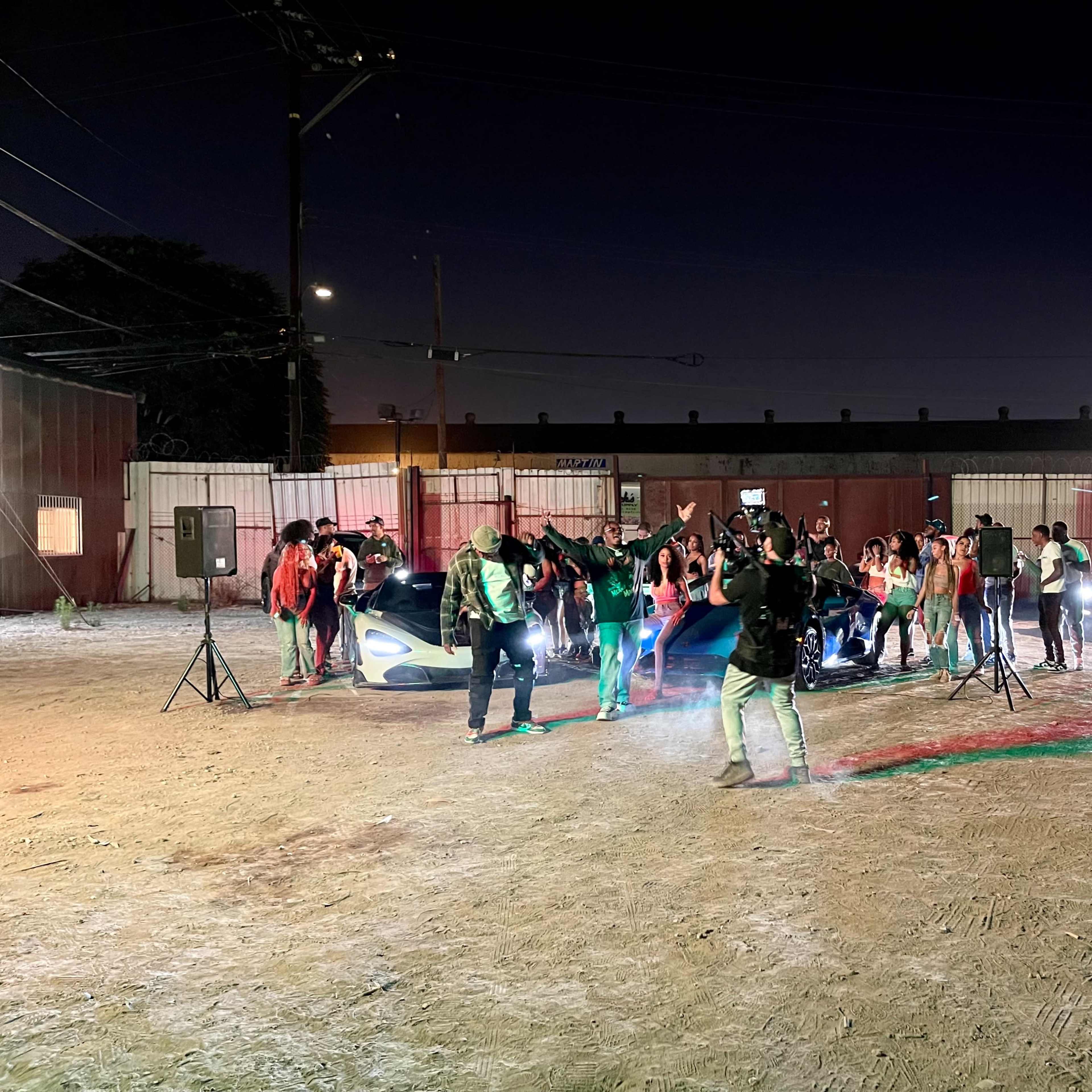 A group of people is gathered around two cars in an outdoor setting at night, with colorful lights illuminating the scene.