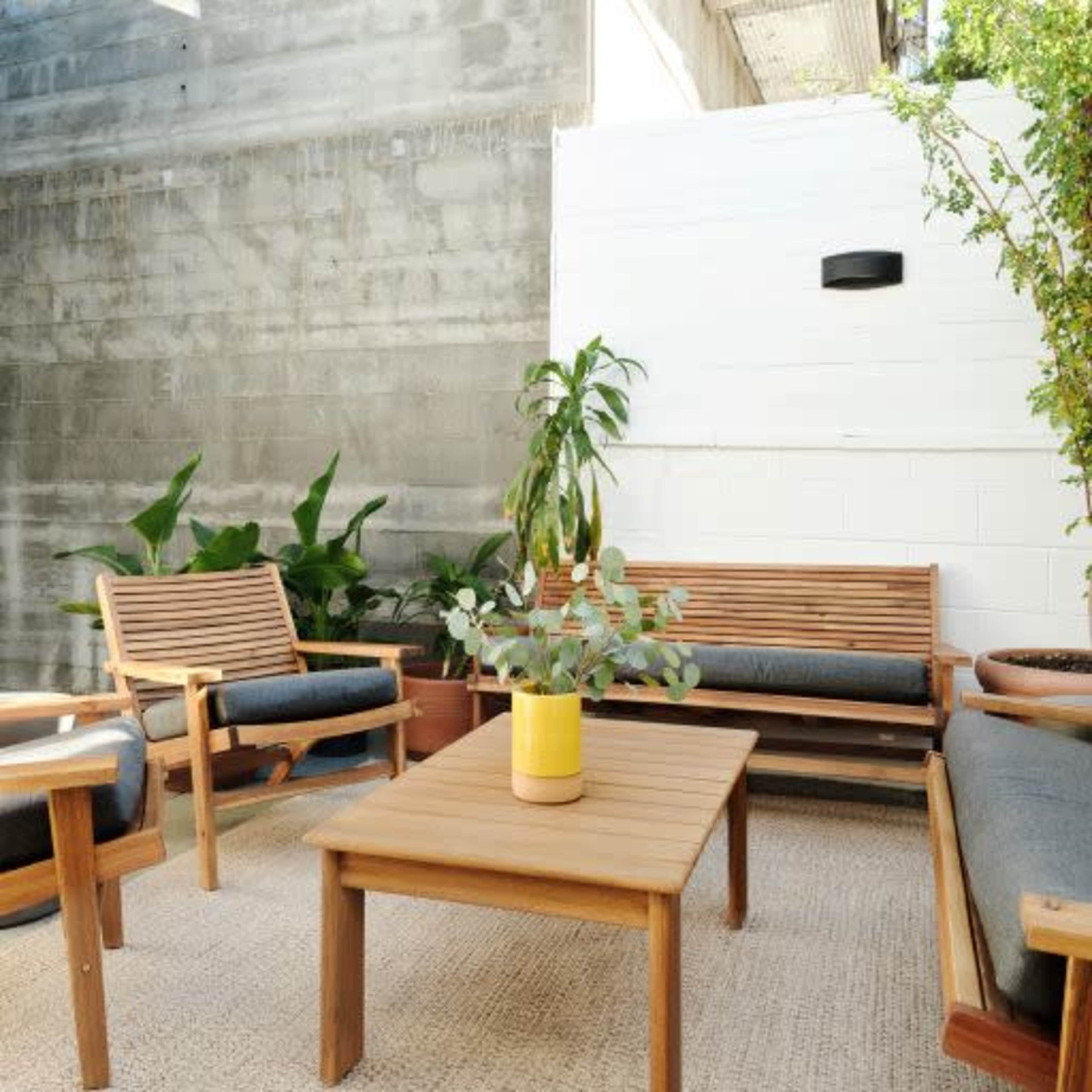 A small outdoor seating area features two wooden chairs and a table surrounded by potted plants against a plain wall.